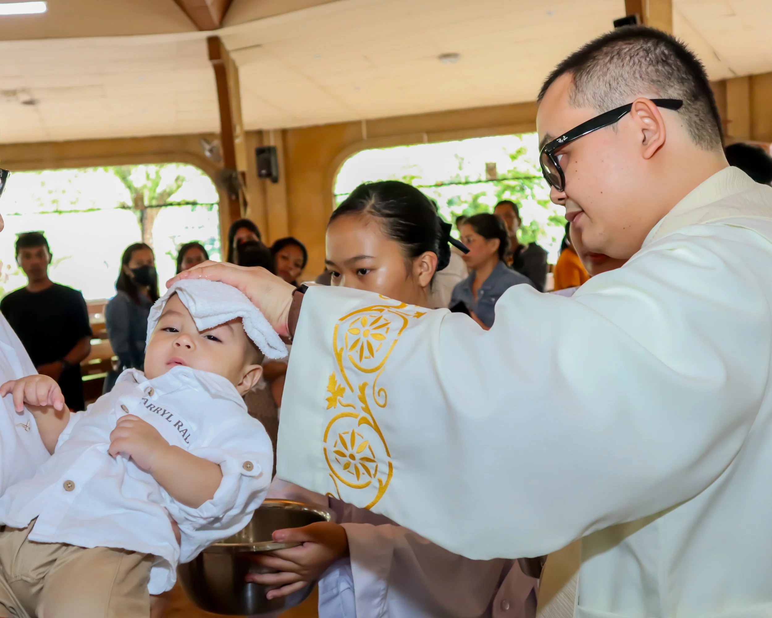 A boy receiving a baptism in a church, with a priest pouring holy water on his forehead while holding a bowl; onlookers are in the background observing the ceremony.
