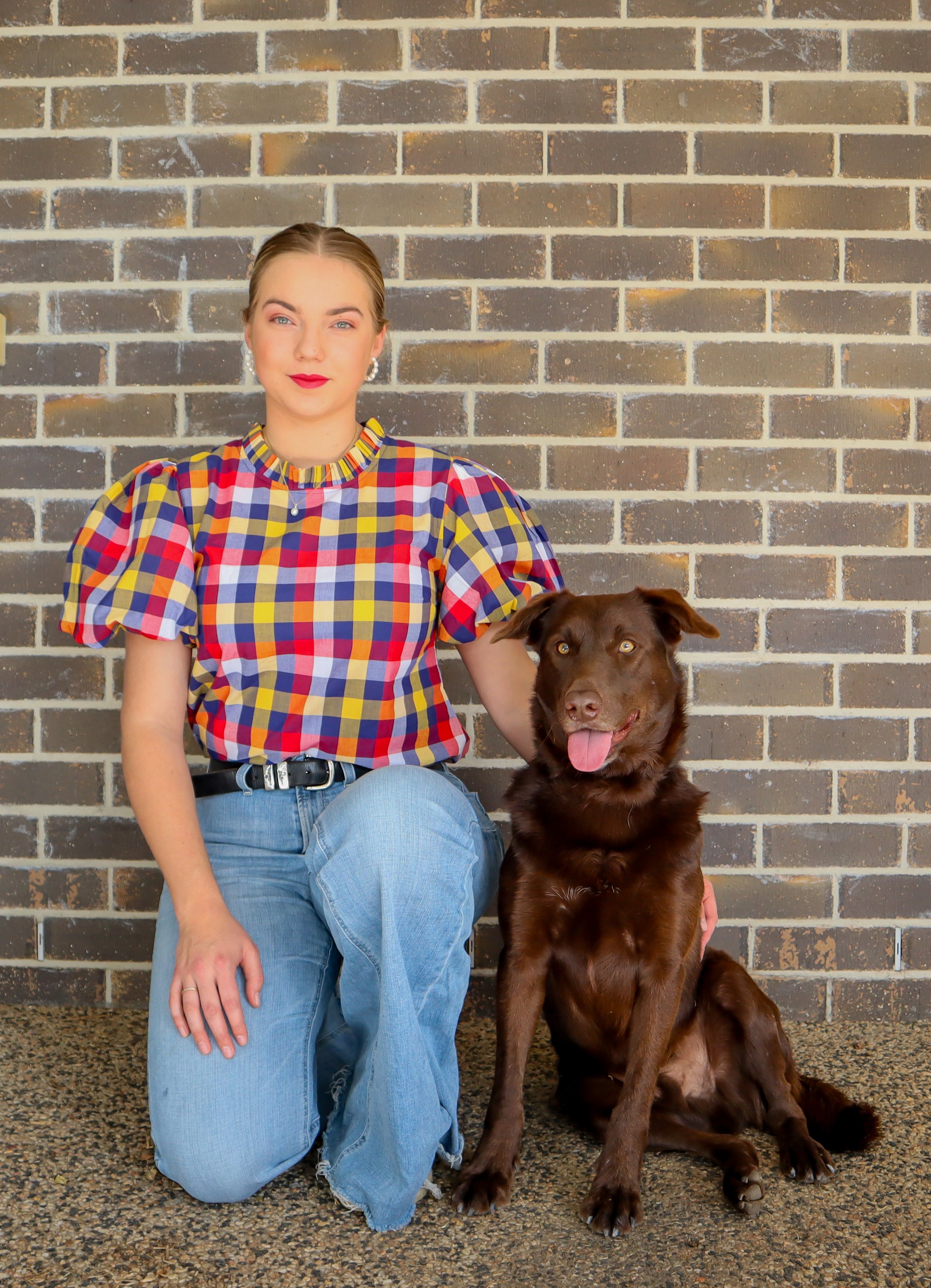 A woman with light skin, blonde hair, and bright red lipstick, wearing a colorful checkered shirt and blue jeans, kneeling next to a sitting brown dog with a pink tongue, in front of a brick wall.