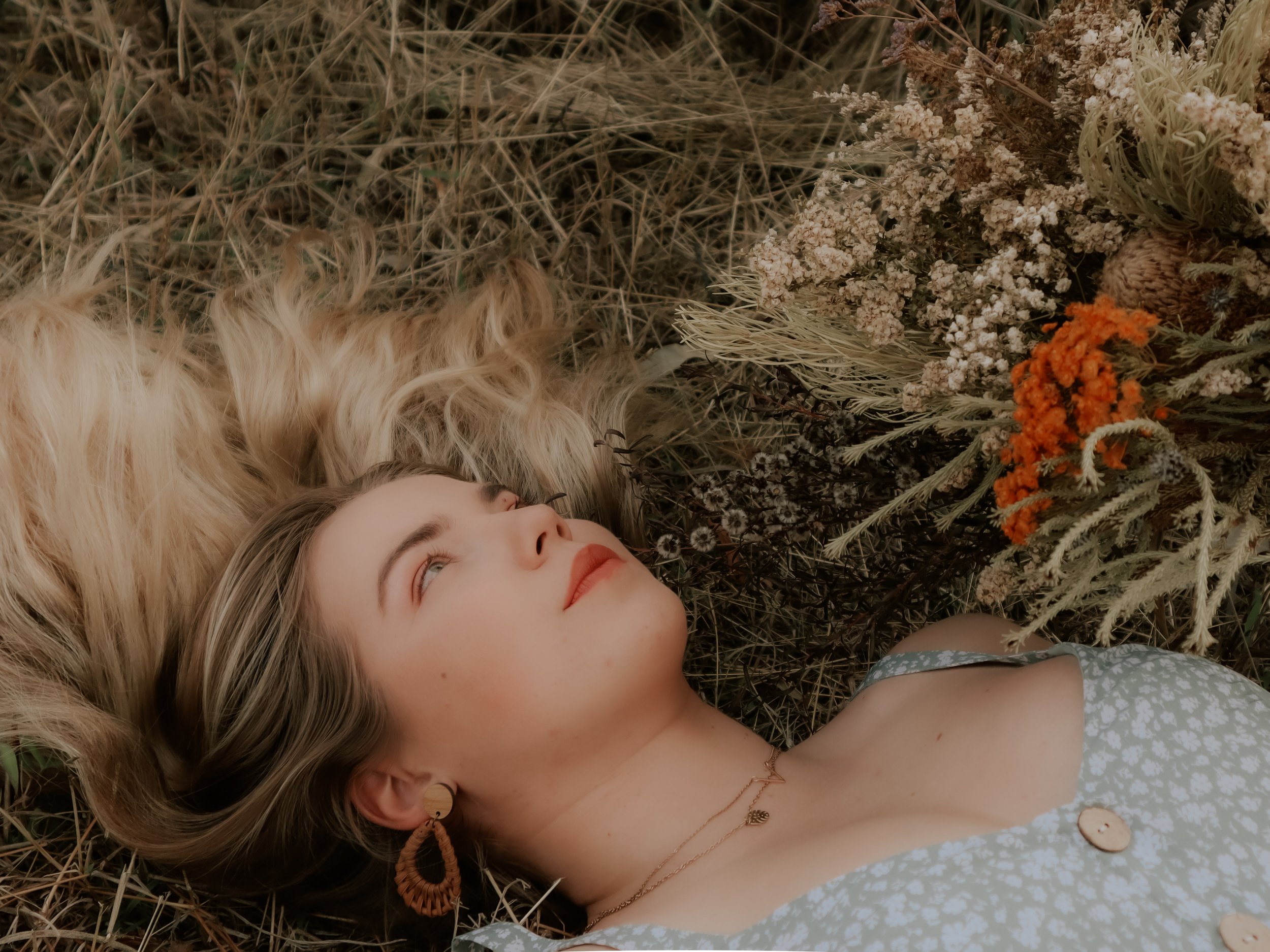 A young woman with blonde hair, earrings, and necklaces lies on the ground surrounded by dried grass and colorful flowers, looking upwards.