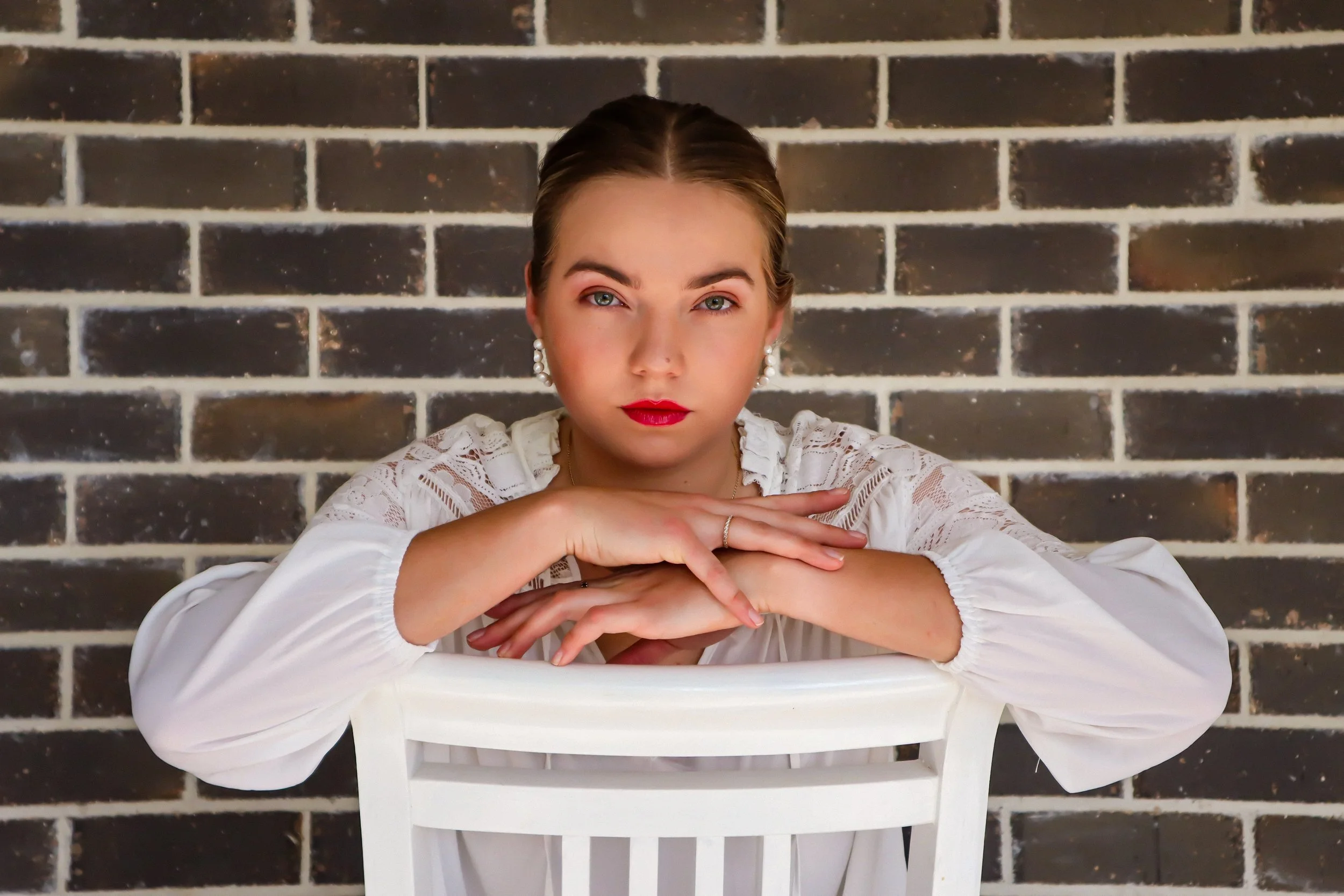 A woman with blonde hair and earrings posing in front of a dark brick wall, resting her chin on her folded arms on the back of a white chair.