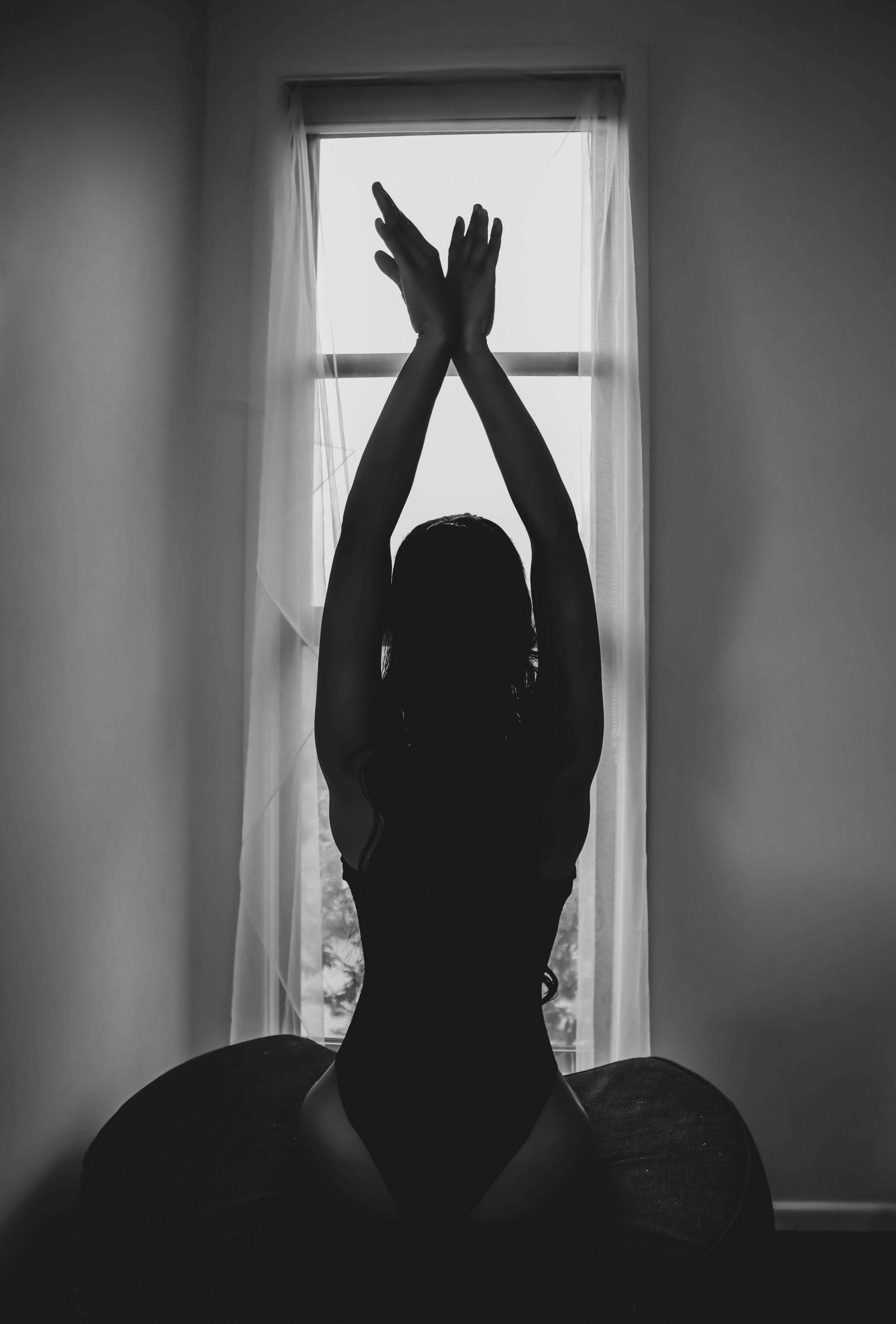 Silhouette of a woman kneeling on a bed with her hands raised above her head, backlit by a window with sheer curtains, in black and white.