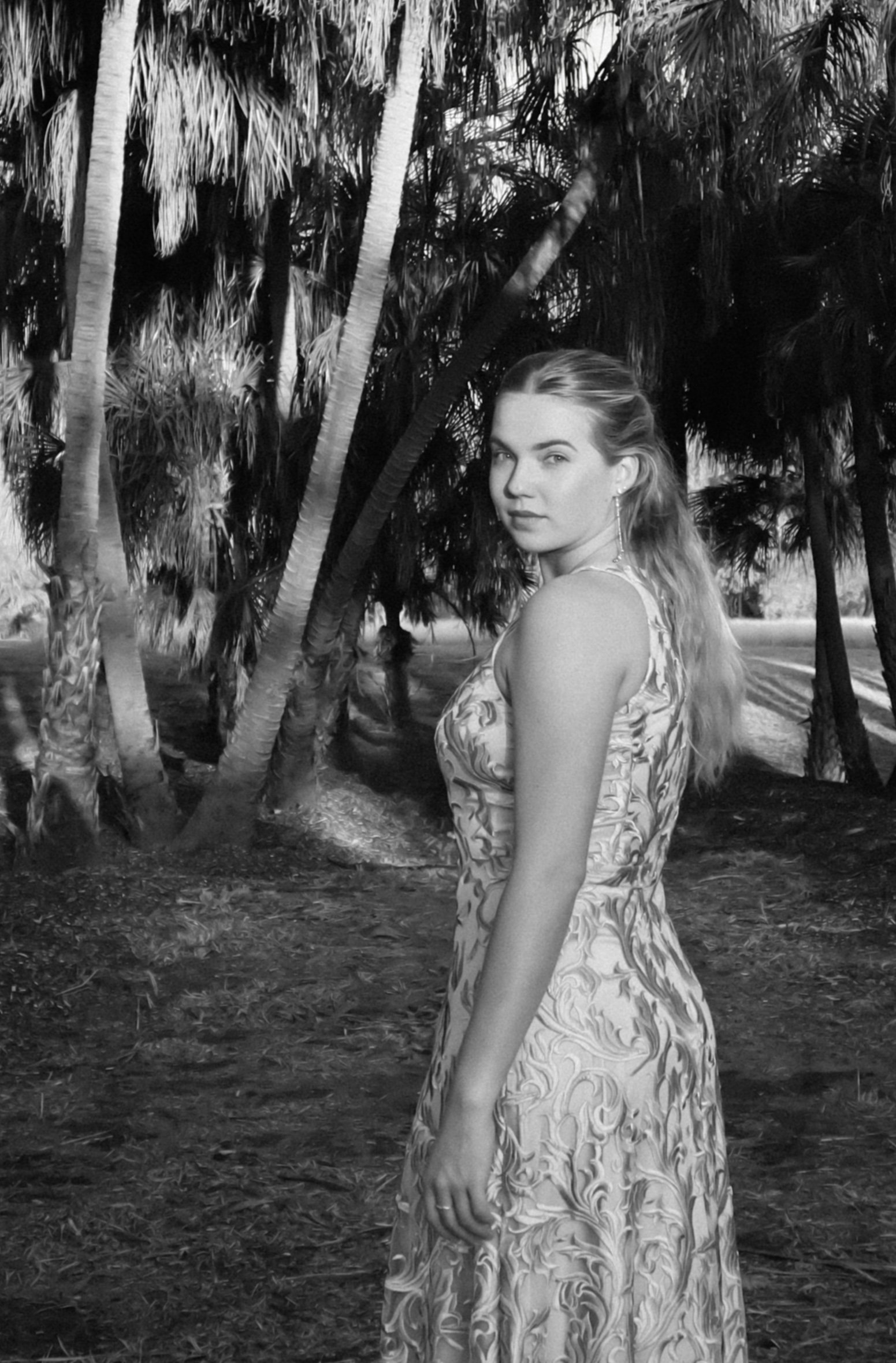 A woman in a patterned dress standing outdoors among palm trees, looking at the camera with a slight smile, in black and white.