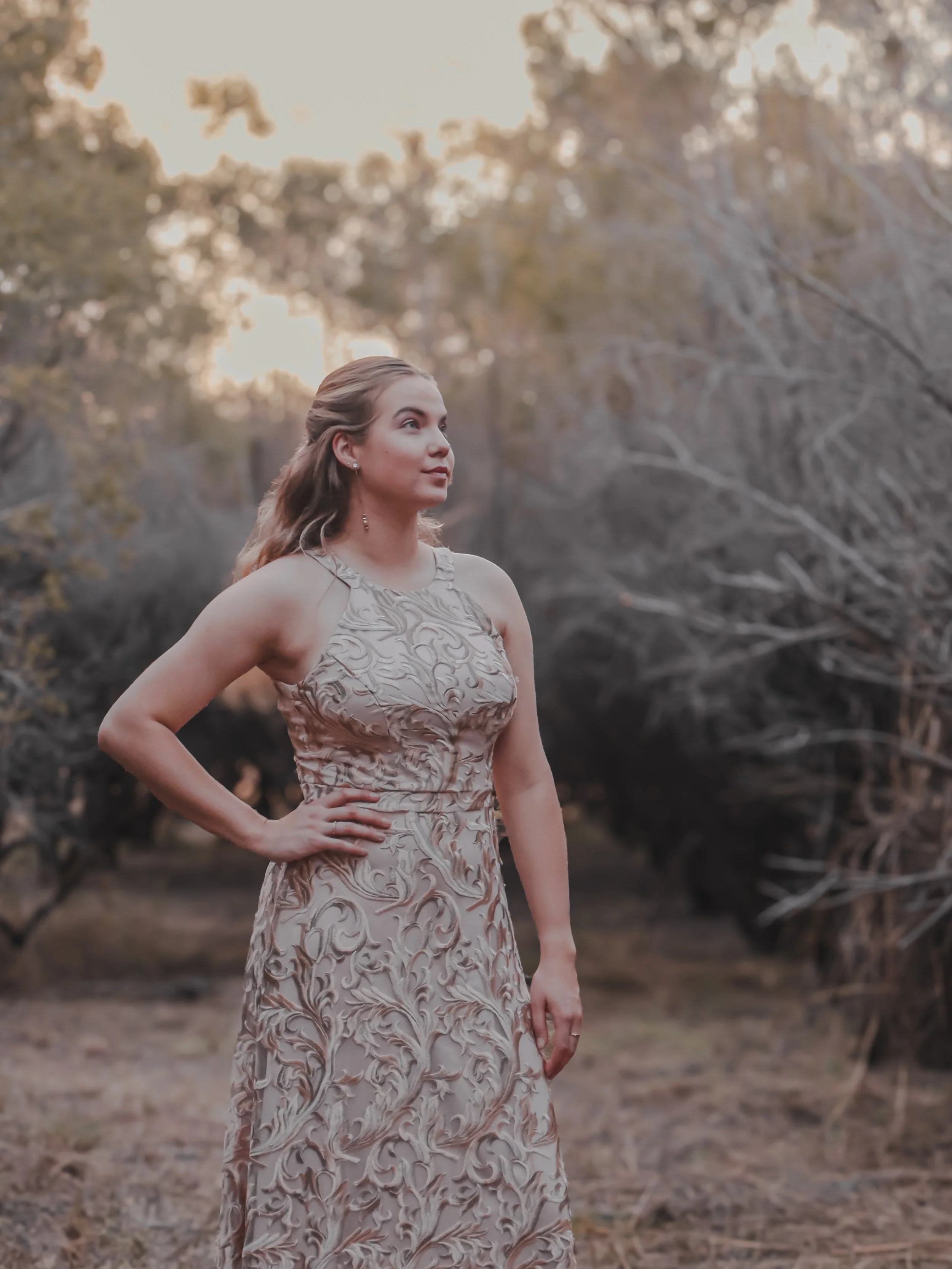 A woman in a beige, embroidered dress stands outdoors with one hand on her hip, looking to her right, in a wooded area during sunset.
