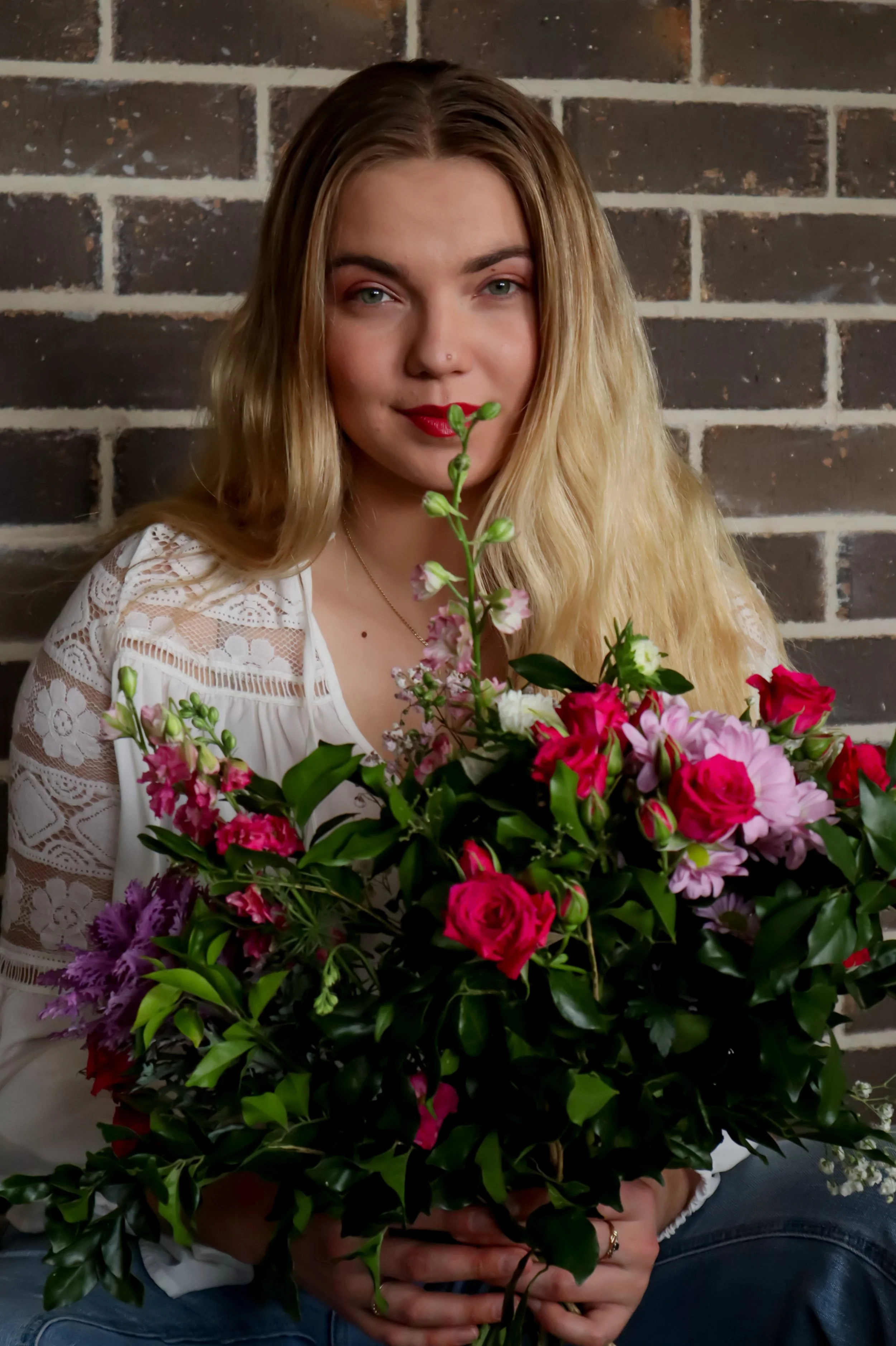 A woman with long blonde hair, blue eyes, and red lipstick holding a large bouquet of colorful flowers in front of a brick wall.
