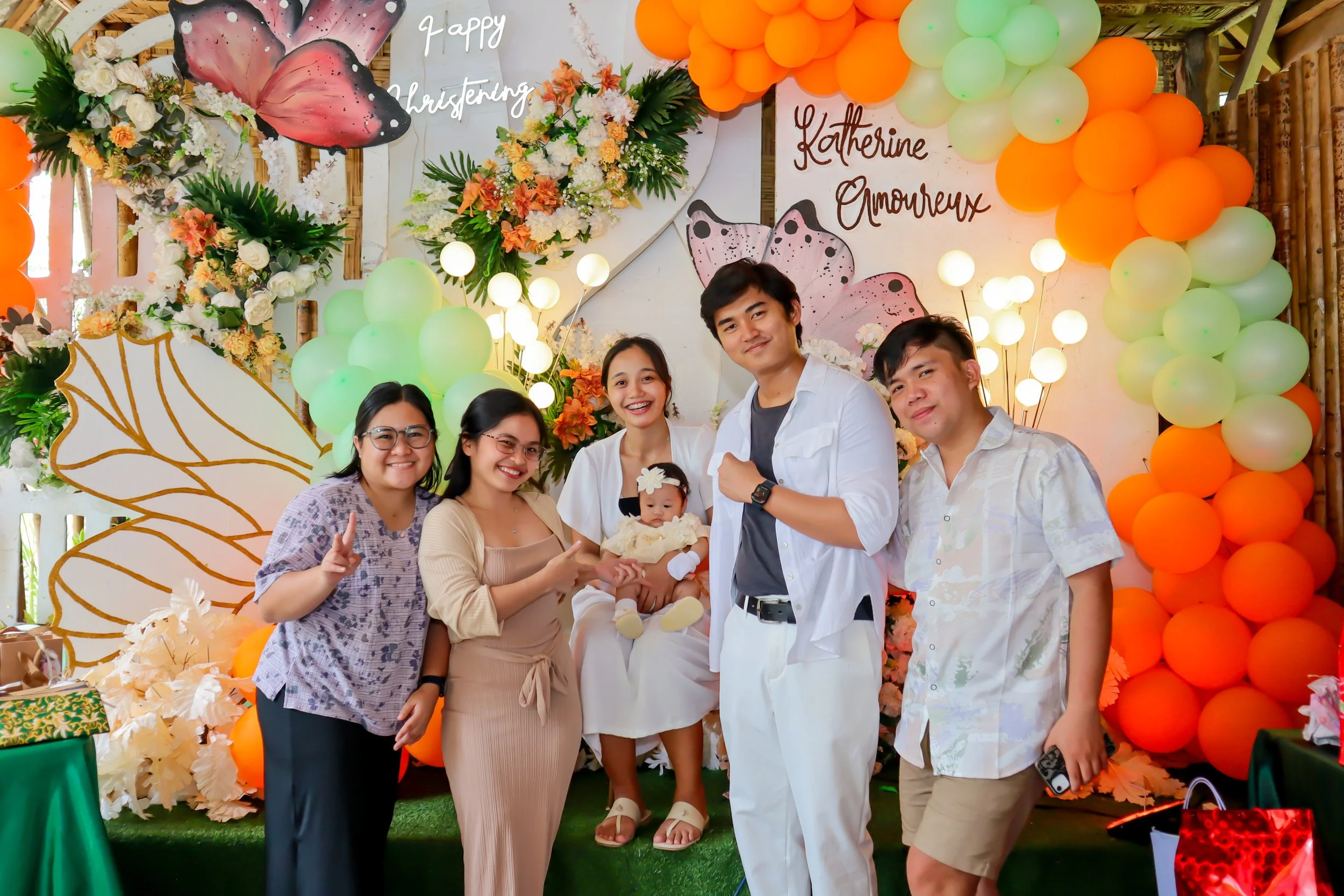 A group of people posing for a photo at a butterfly-themed celebration with floral decorations and balloons. The backdrop reads "Happy Chrystening" and "Katherine Amoureux."
