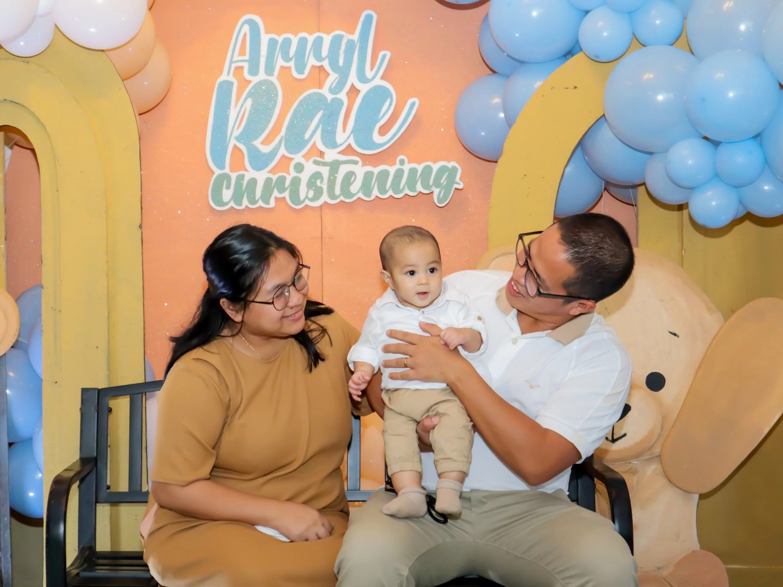 A family of three sitting on a bench at a Christening celebration, with colorful balloons and a yellow and pink backdrop that says 'Arrival RAE Christening'. The father is holding a young child on his lap, and the mother is sitting beside them, smili