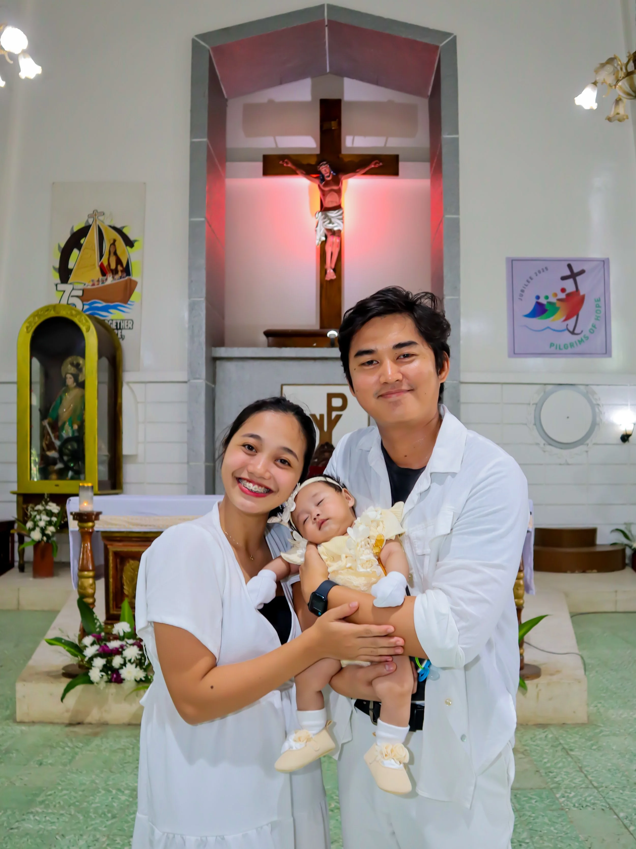 A family of three in a church after a baptism ceremony, with a crucifix and religious symbols in the background. The parents are holding their baby daughter, who is dressed in yellow and white.