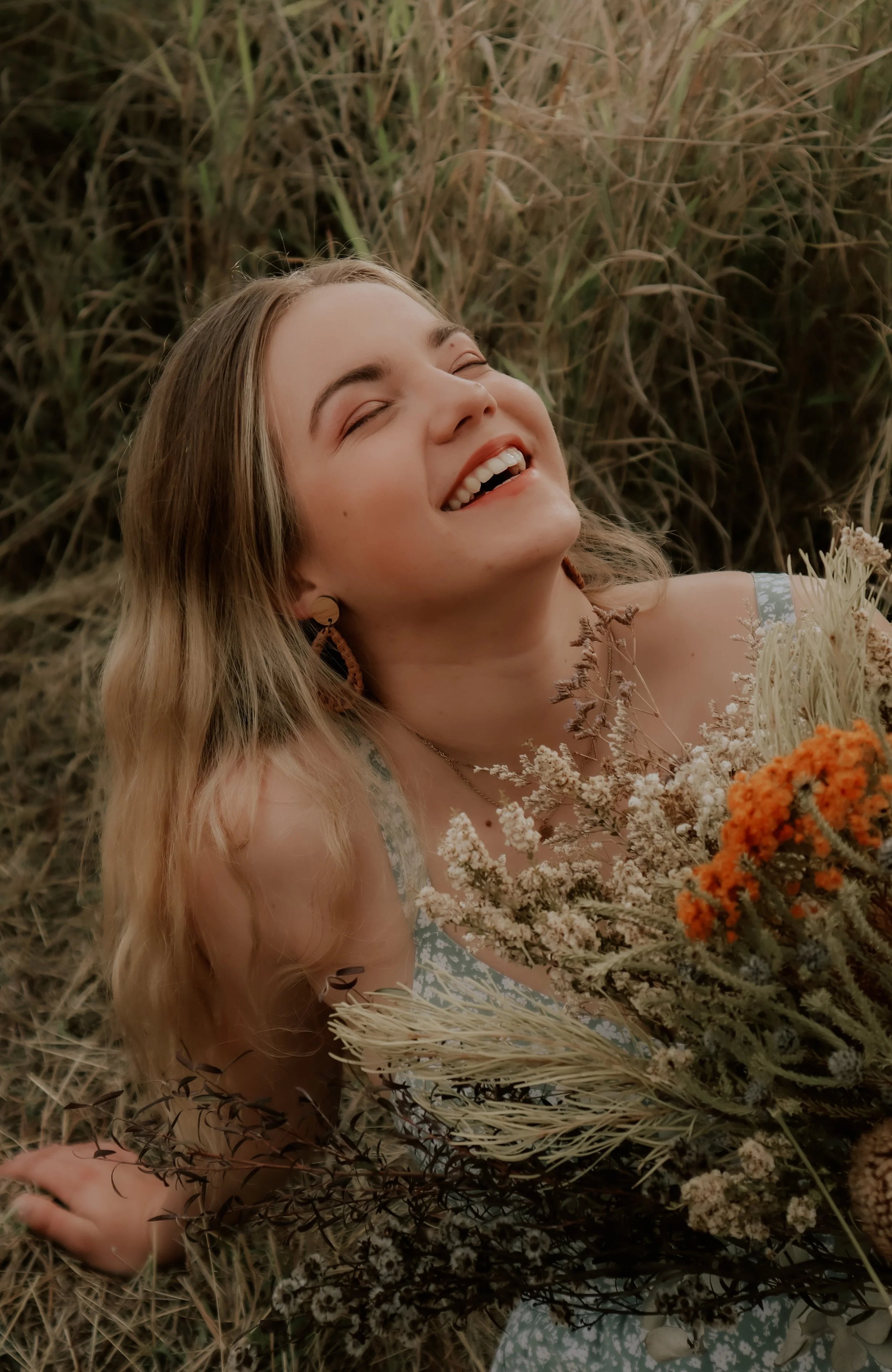 A smiling woman with long blonde hair lying down in a field of tall grass, holding a bouquet of dried flowers.