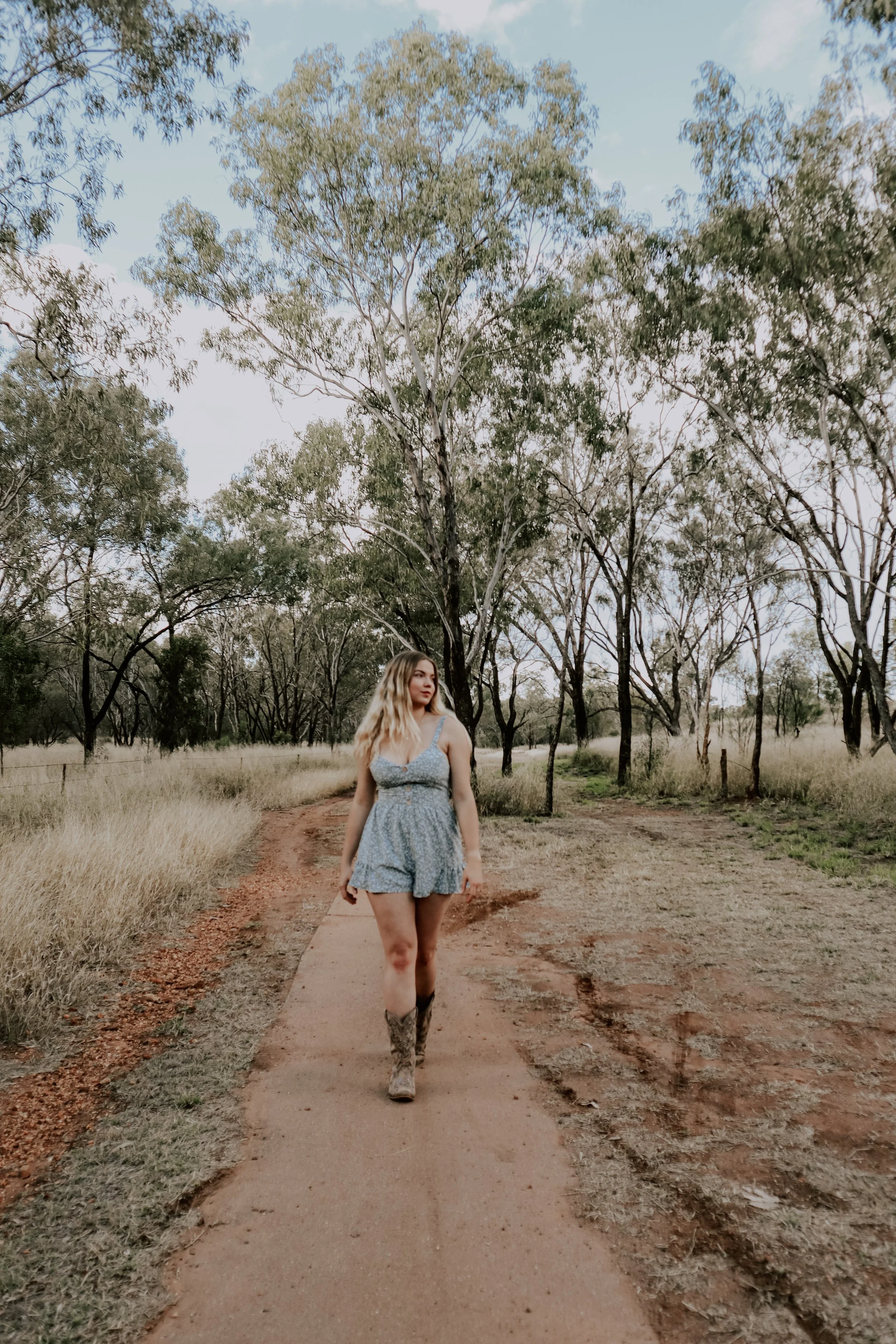 A woman walking a dirt path in a rural area with trees and tall grass on both sides.