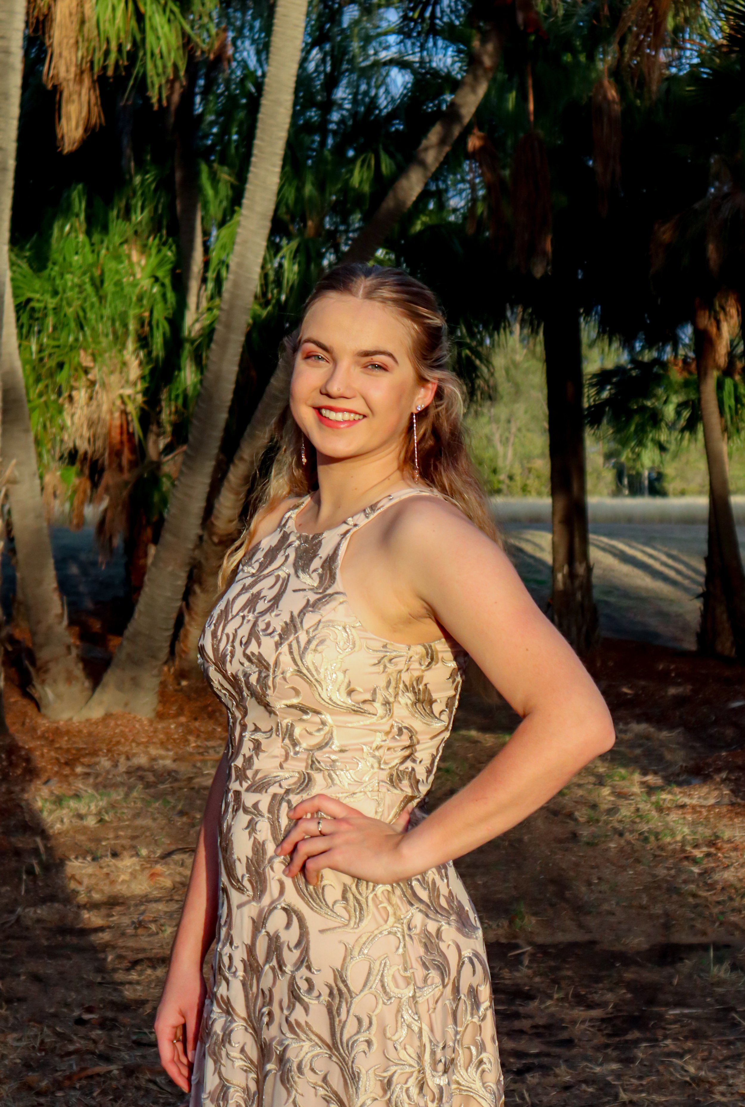 A young woman in a beige and gold patterned gown standing outdoors with her hand on her hip, smiling at the camera, surrounded by palm trees and sunny weather.