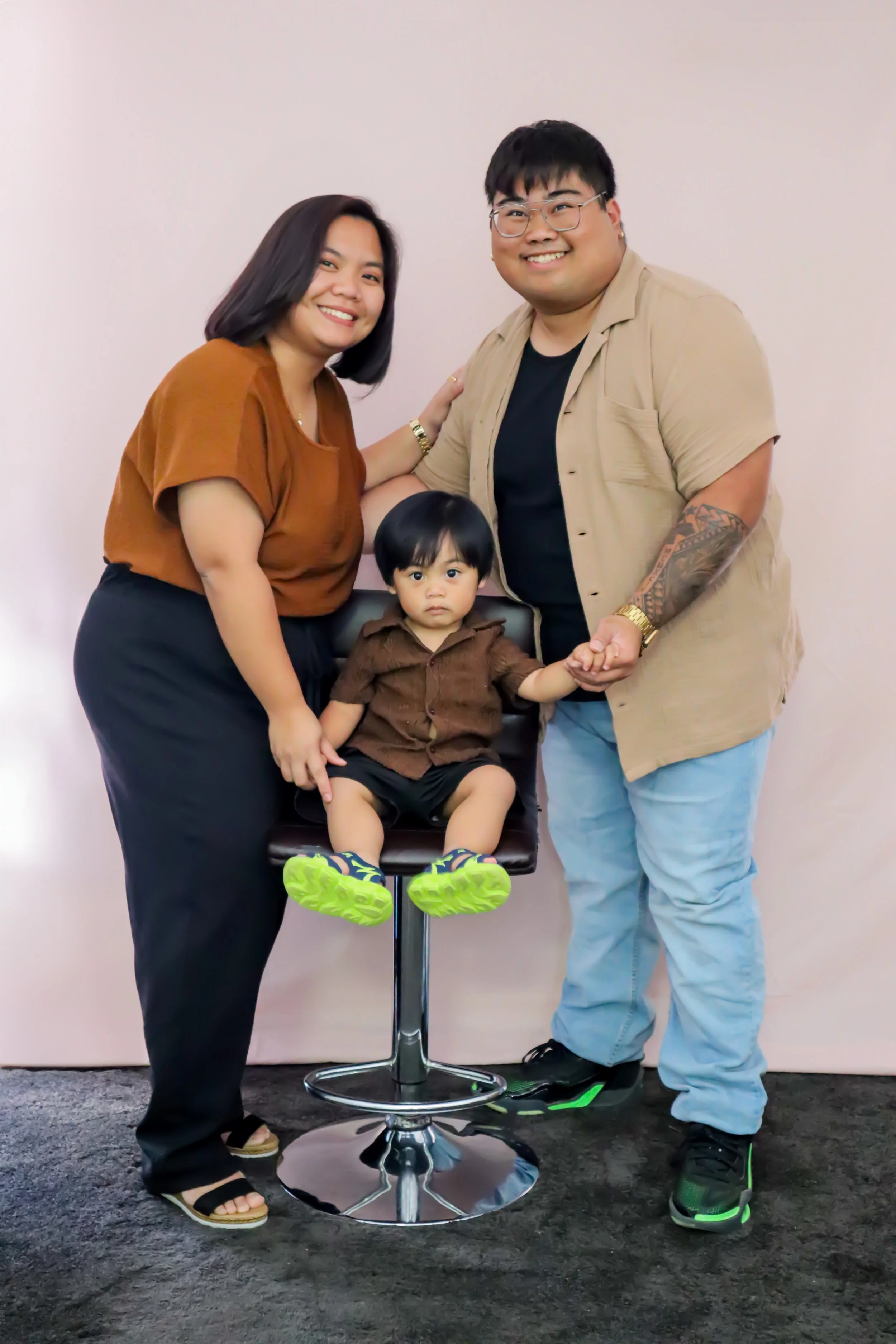 Family portrait of three people and a young boy, standing in front of a plain white background. The woman is on the left wearing a brown top and black pants, smiling. The man is on the right wearing glasses, a beige shirt, and light blue pants, also 