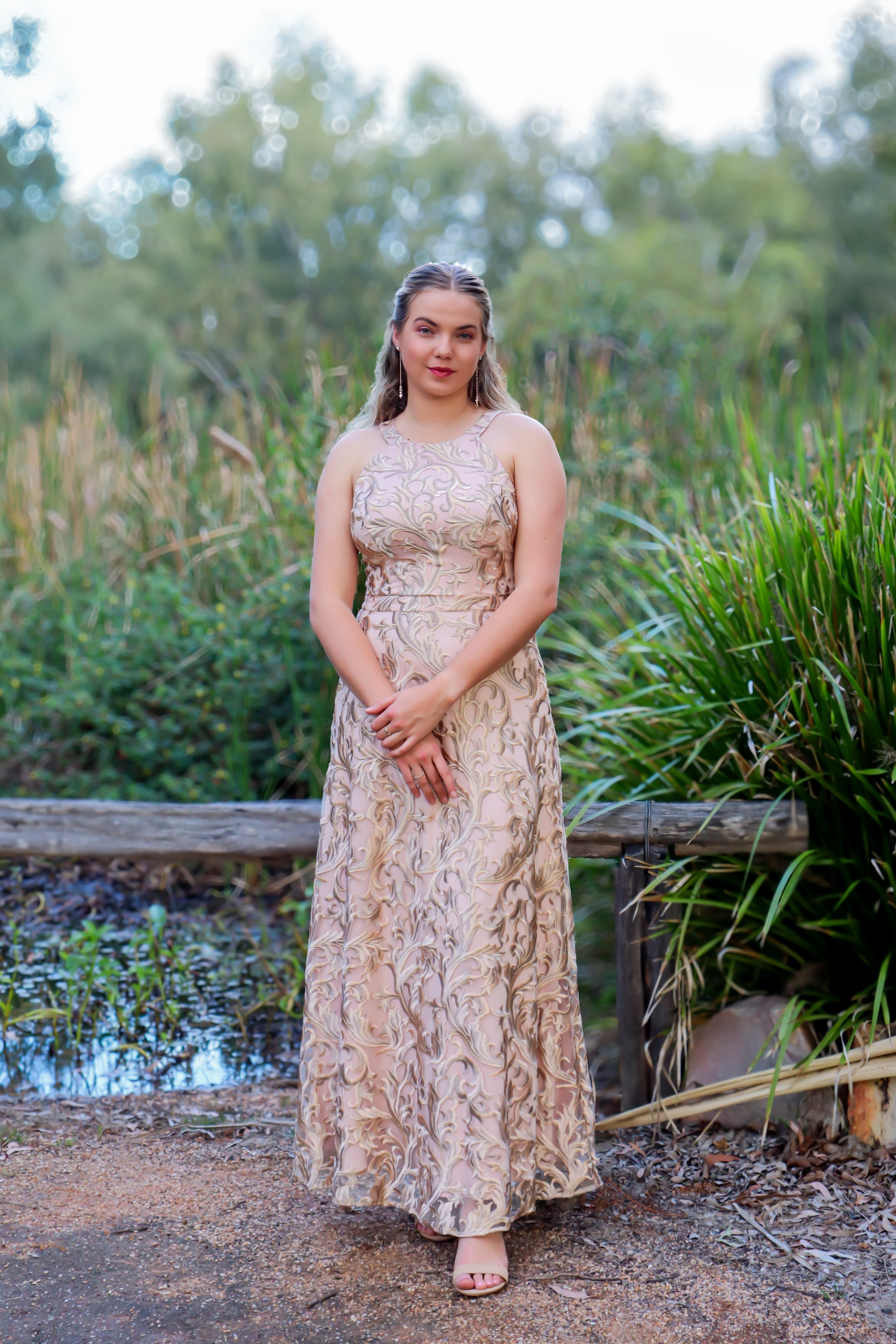 Young woman in a long, intricate beige and gold dress standing outdoors near greenery and a small pond.