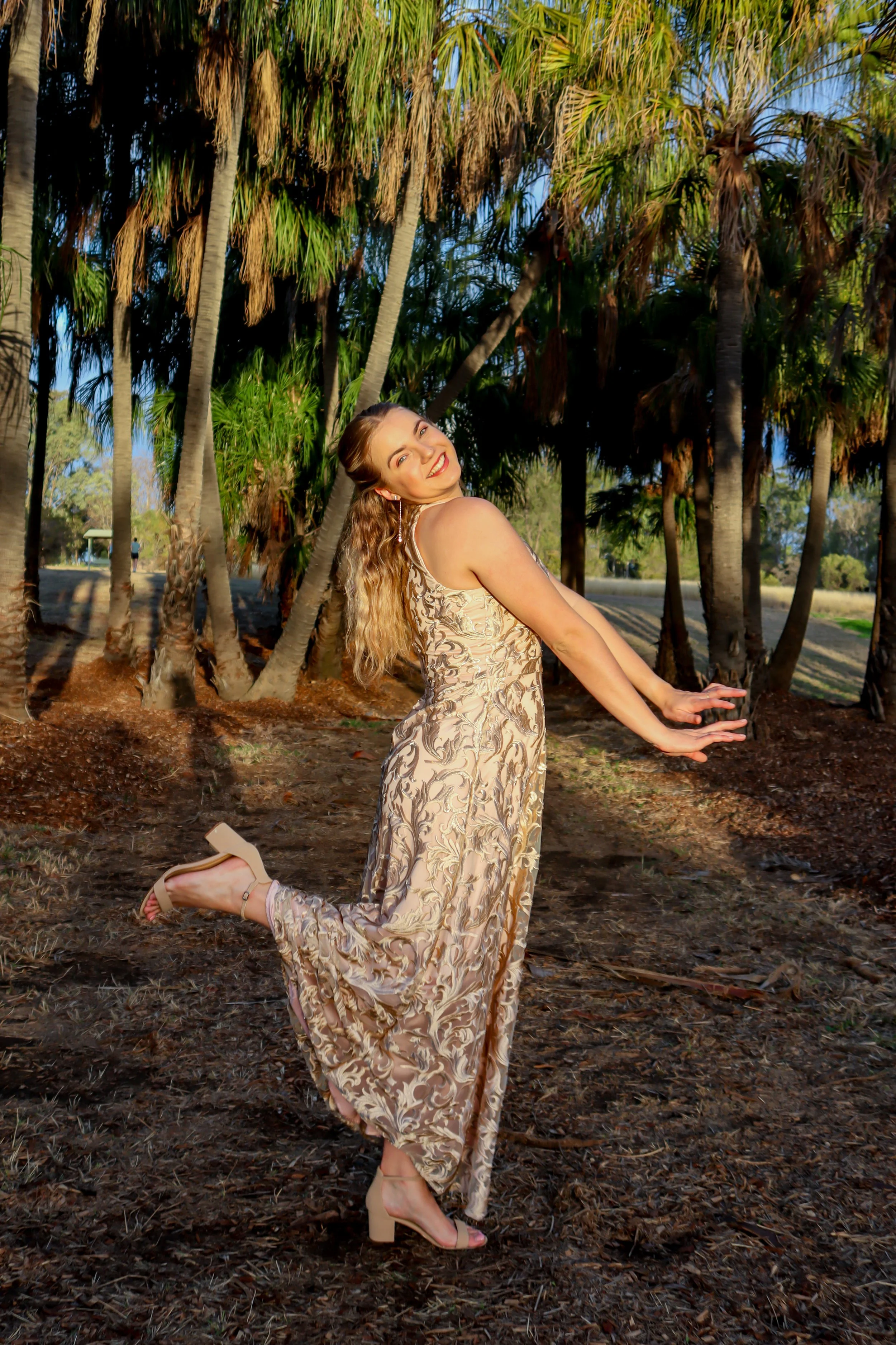 A woman with long, wavy blonde hair is standing in a sunlit forest of tall palm trees, wearing a flowing patterned gown and beige heels, smiling and posing with one leg lifted and arms extended.