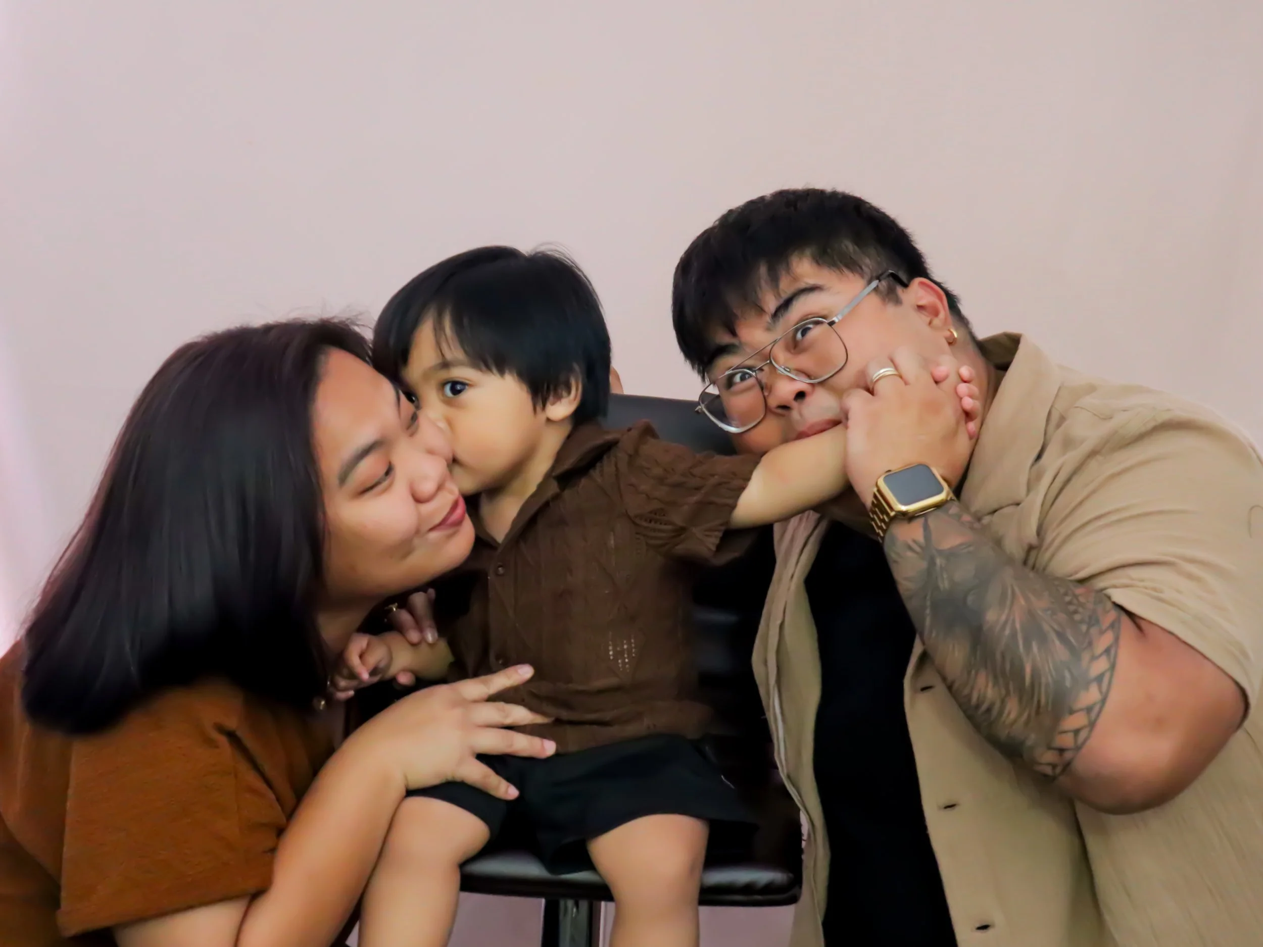A family of three, including a woman, a young boy, and a man, making playful faces and gestures against a plain light-colored wall.