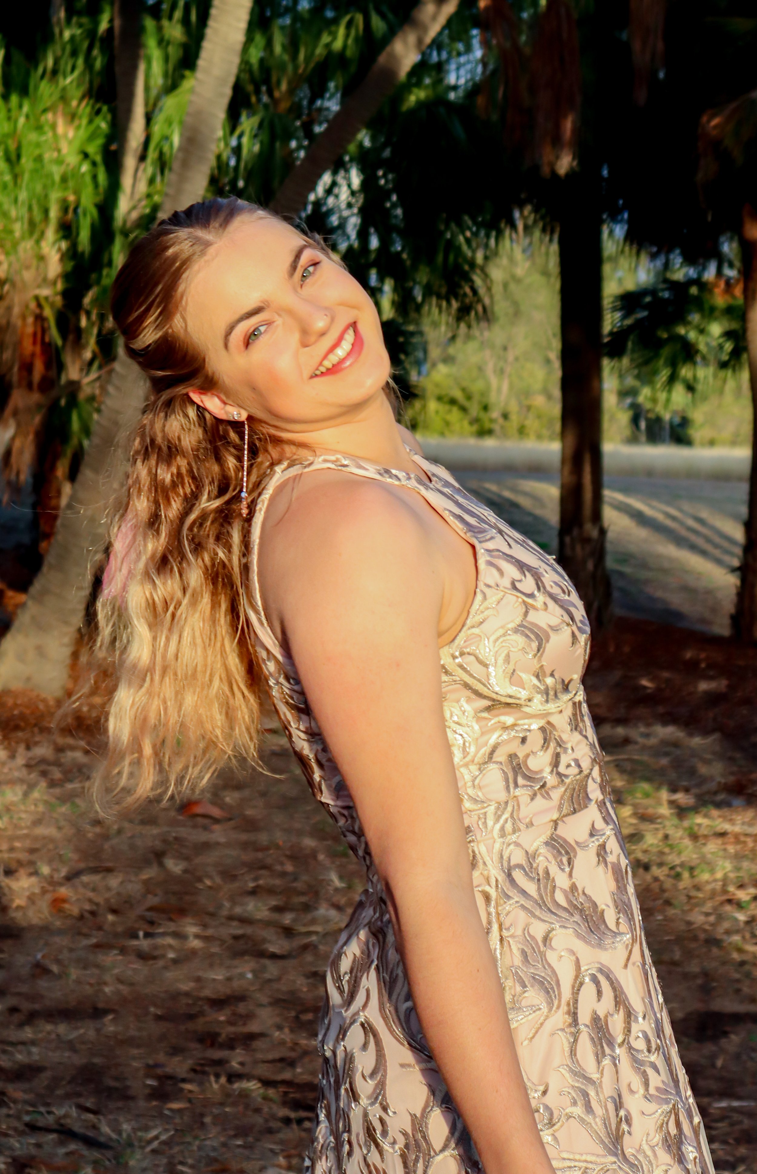 A young woman with long, wavy blonde hair, smiling and looking over her shoulder outdoors in a sunny, wooded area.