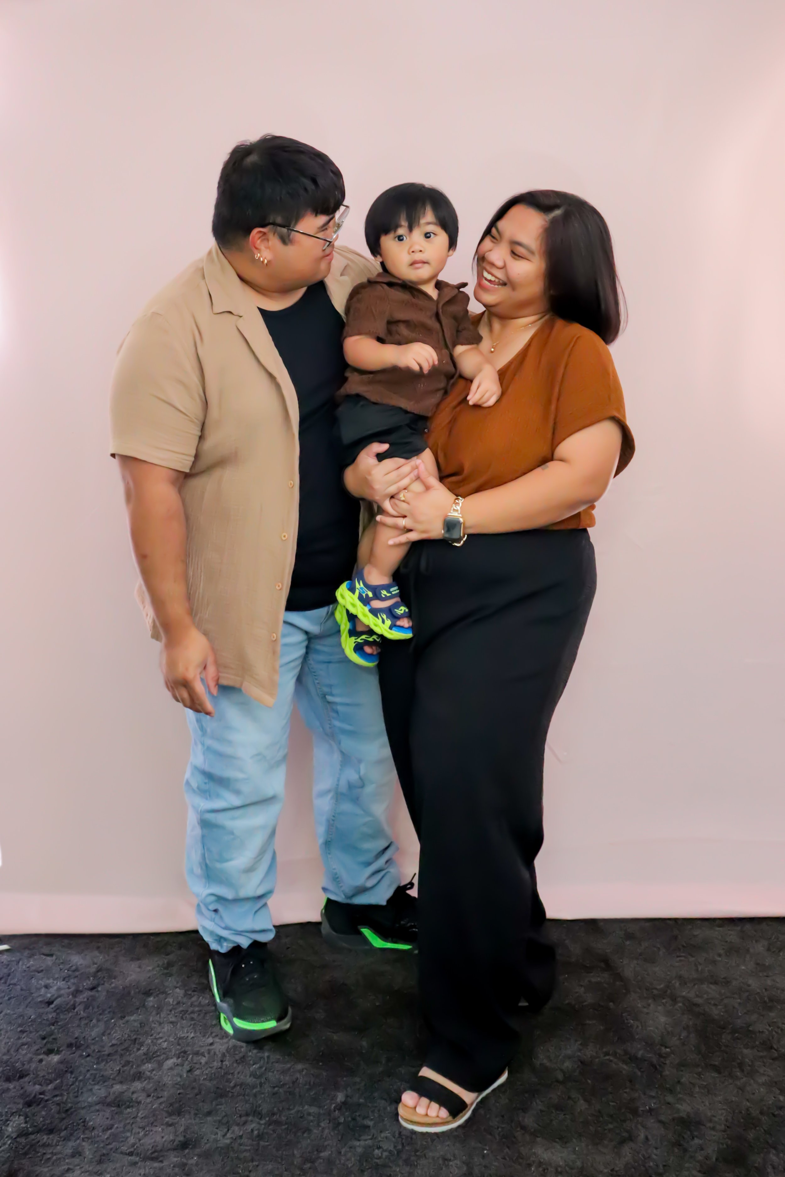 Three people, a man, a woman, and a young boy, standing together against a plain wall. The man on the left is wearing glasses, a beige shirt, and light-colored jeans. The woman on the right is wearing a brown top and black pants, holding the boy in h
