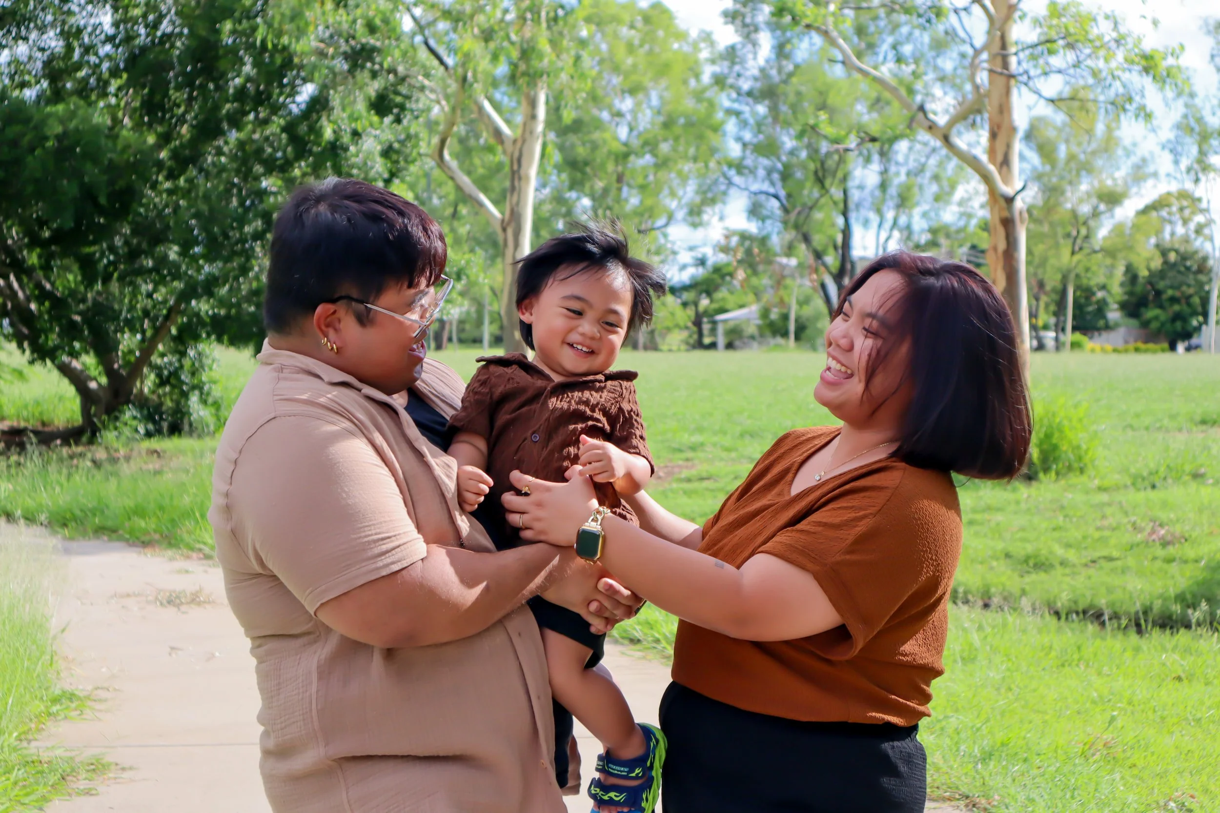 Two adults and a young child outdoors in a park, smiling and playing together, with trees and grass in the background.