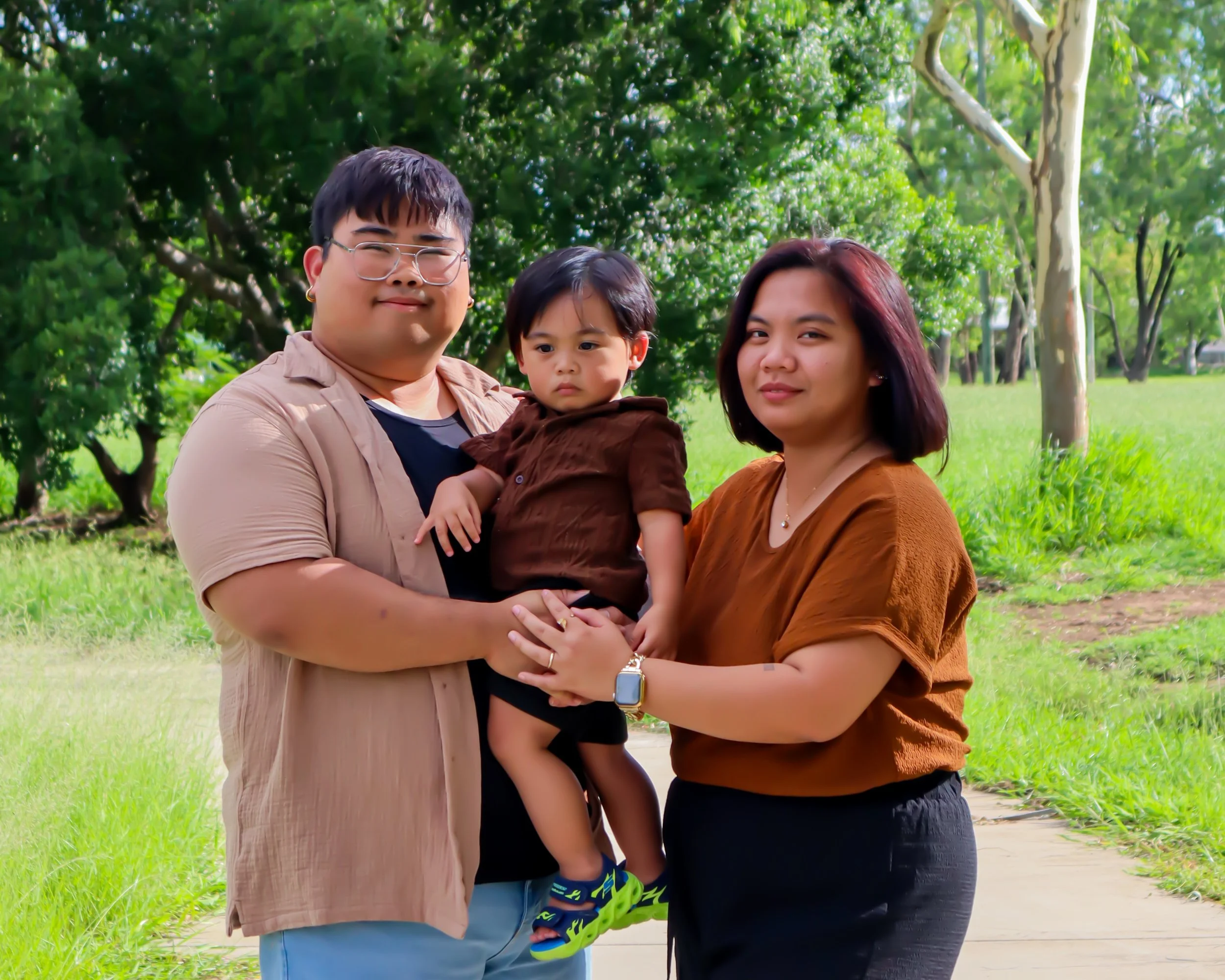 A family photo of two adults and a young boy outdoors in a park with green trees and grass. The man is holding the boy, and the woman is standing next to them. All are smiling and facing the camera.