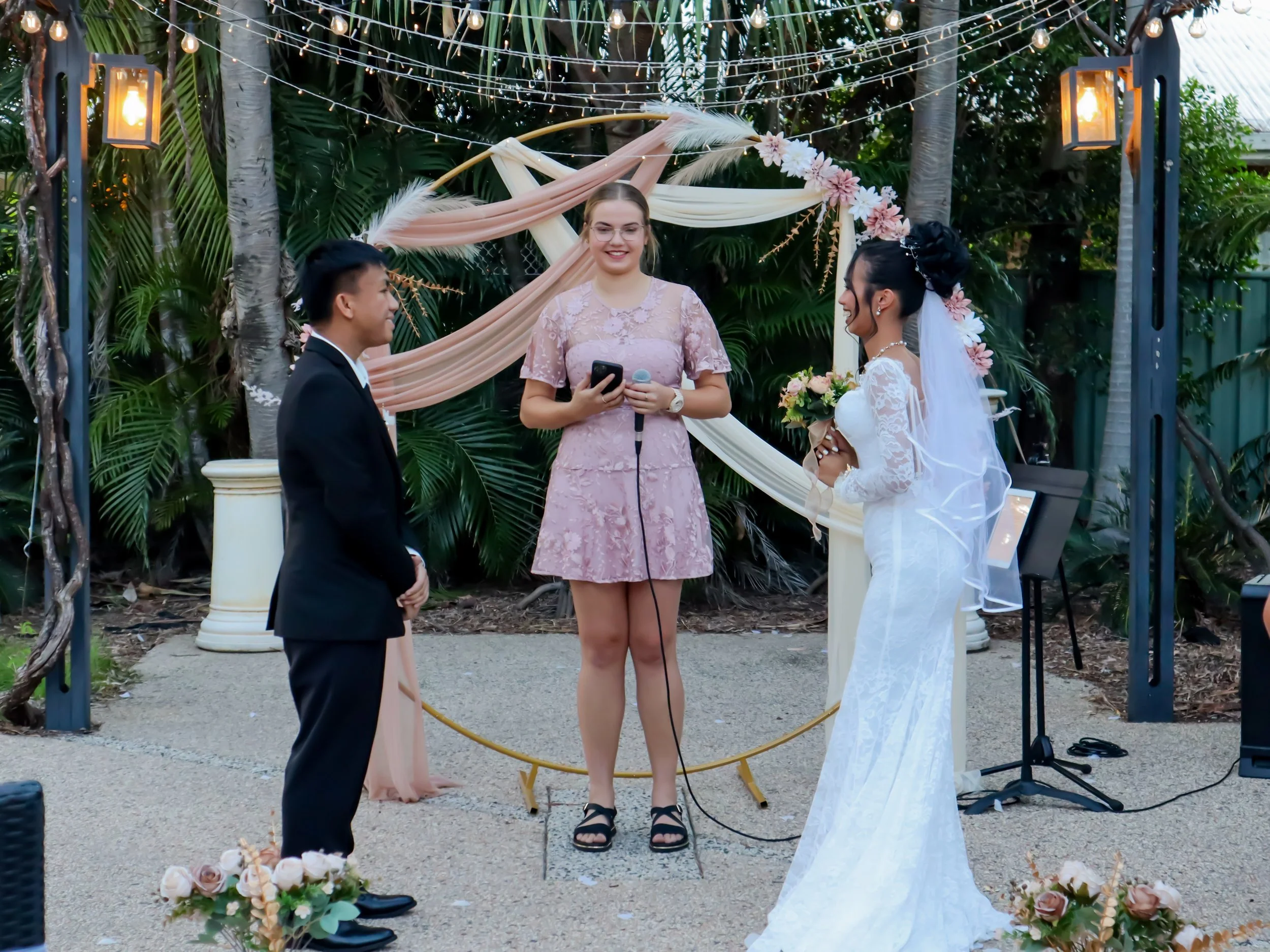A couple getting married outdoors under string lights and floral decorations, with an officiant in pink holding a microphone, as they exchange vows and look at each other.