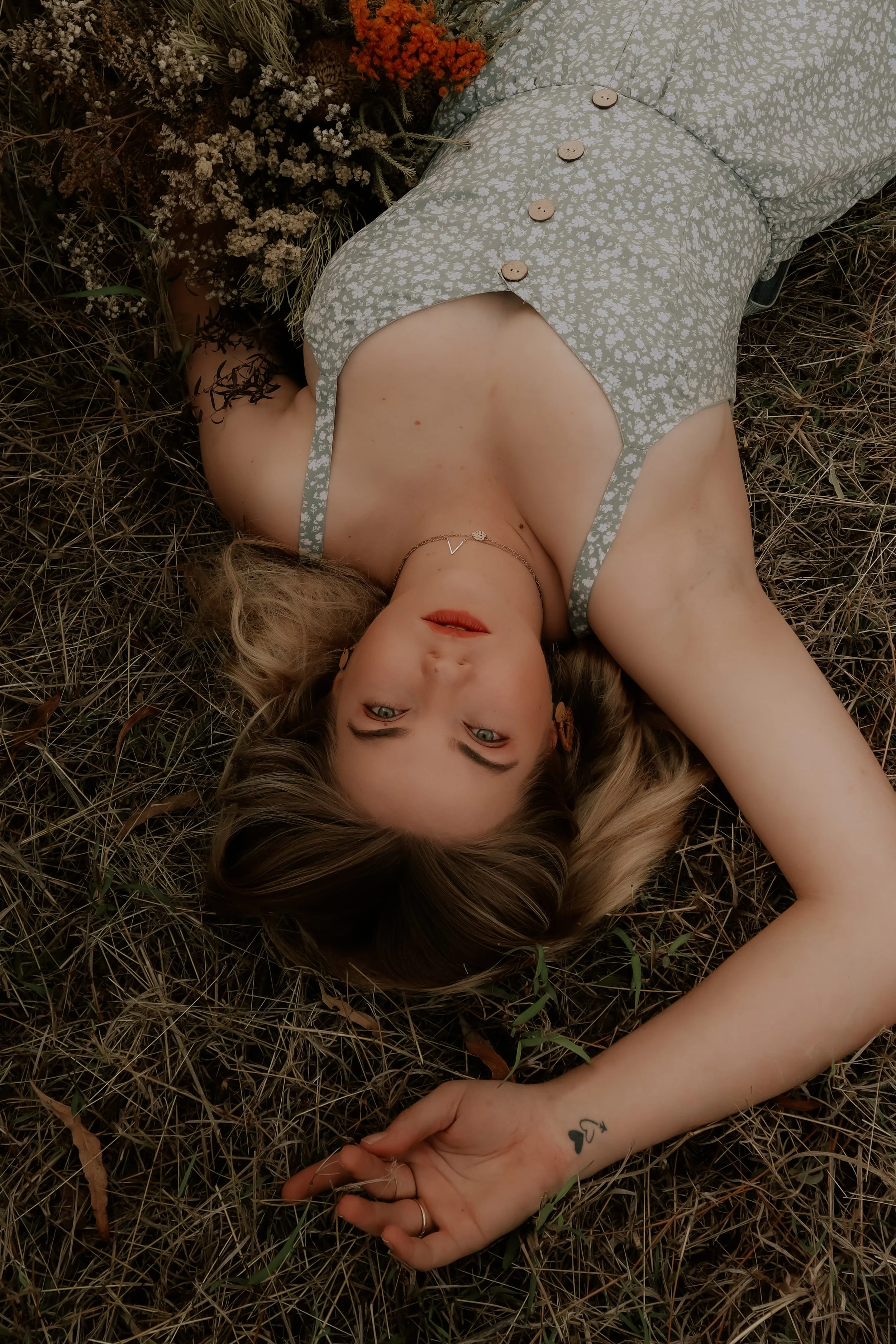 A woman lying on her back on dry grass, wearing a floral dress with buttons, with her head turned to the side and her eyes open. She has blonde hair, a necklace, earrings, and a tattoo on her left wrist.