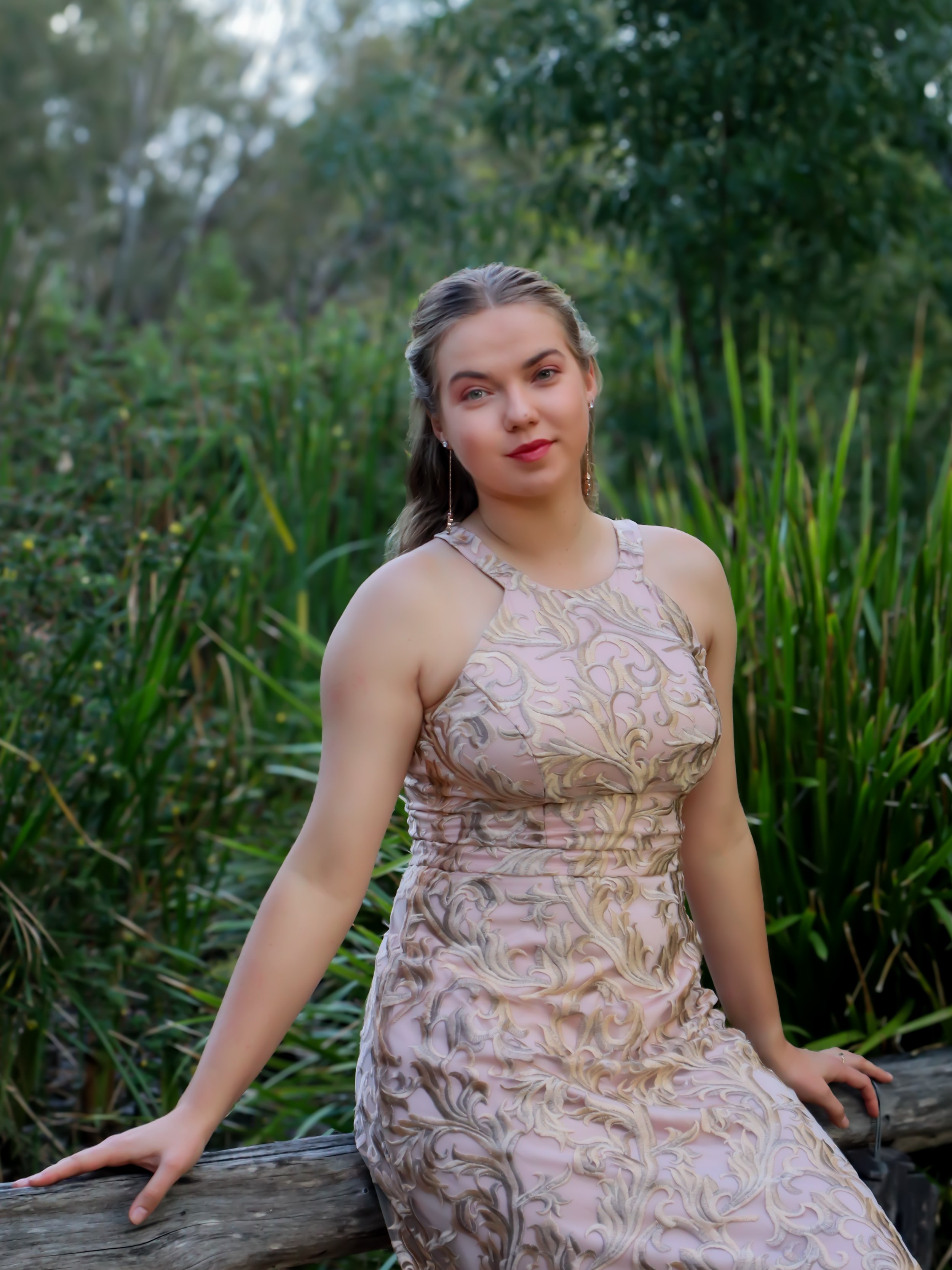 A young woman in a light pink, embroidered dress sits on a log in a lush, green outdoor setting.