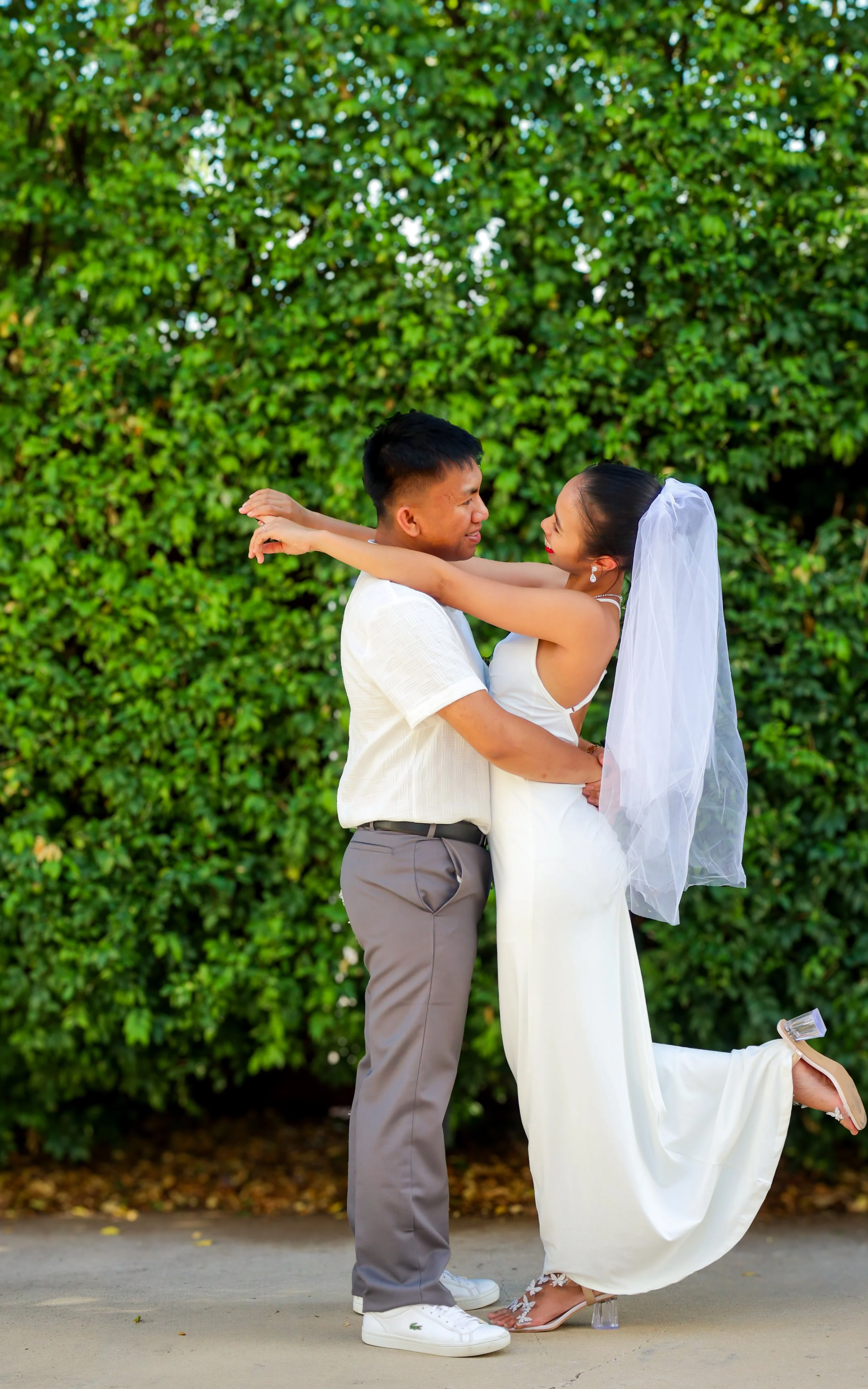 A couple, dressed in wedding attire, embracing and smiling outdoors in front of a lush green hedge.
