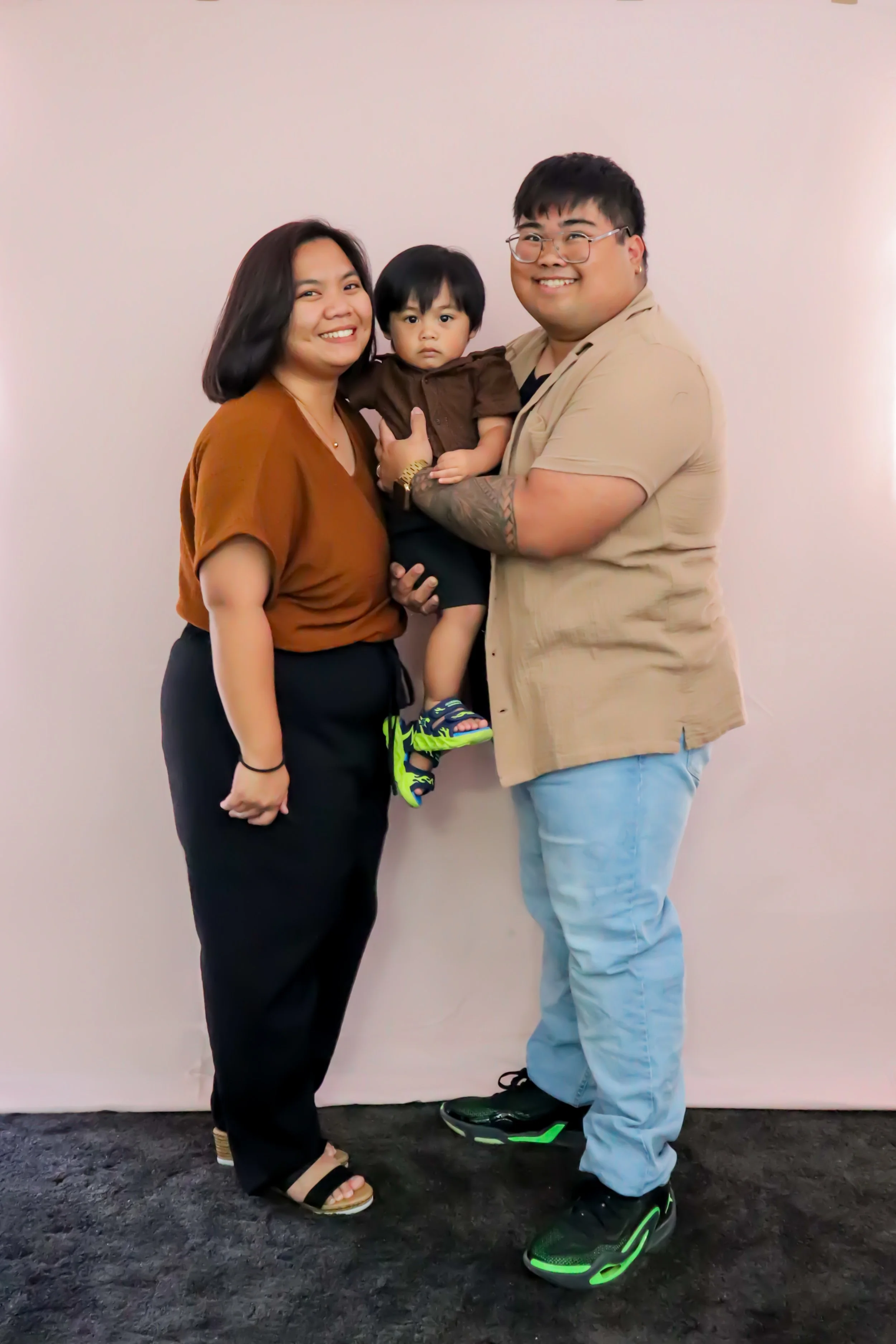 A smiling family of three posing together against a plain wall. The mother has short dark hair and is wearing a brown top and black pants. The father, wearing glasses and a tan shirt, is holding a young boy dressed in a brown T-shirt and black shorts