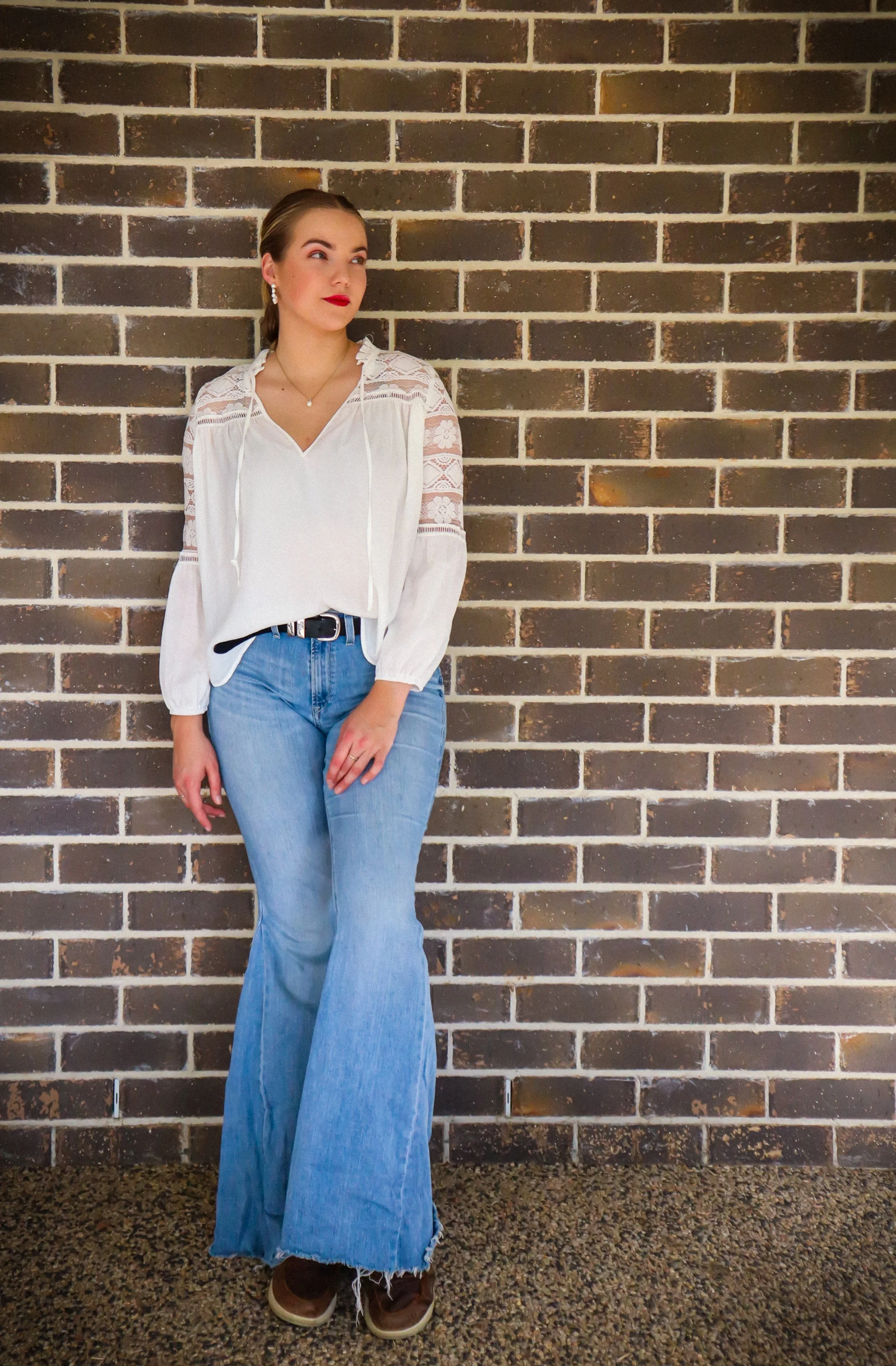 Young woman wearing a white lace shirt, blue jeans, and brown shoes, standing against a brick wall with her hair pulled back and looking to the side.