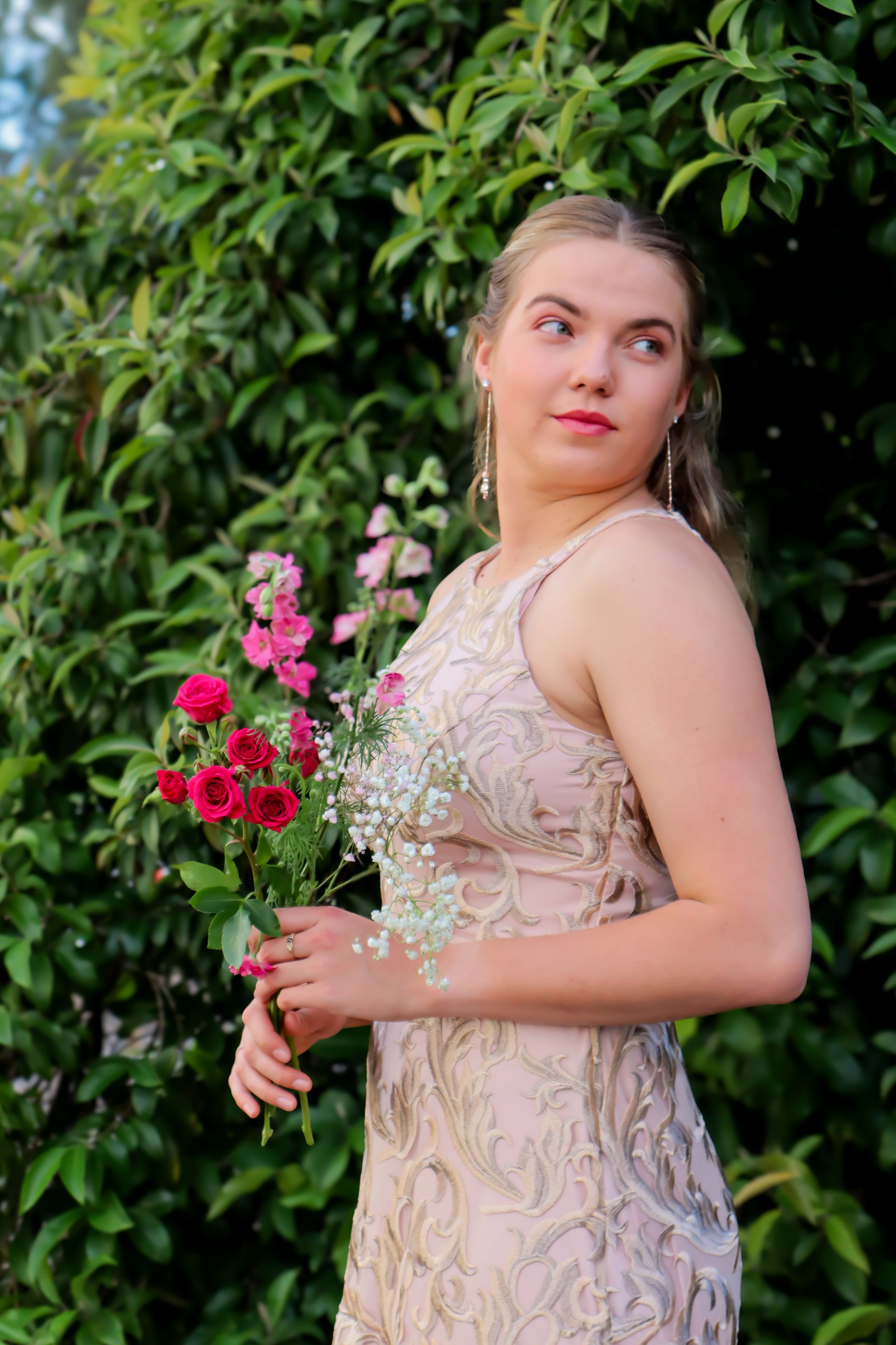 A young woman with blonde hair in an elegant dress holding a bouquet of pink and red roses and greenery, standing outdoors in front of a lush green bush, looking to her right.