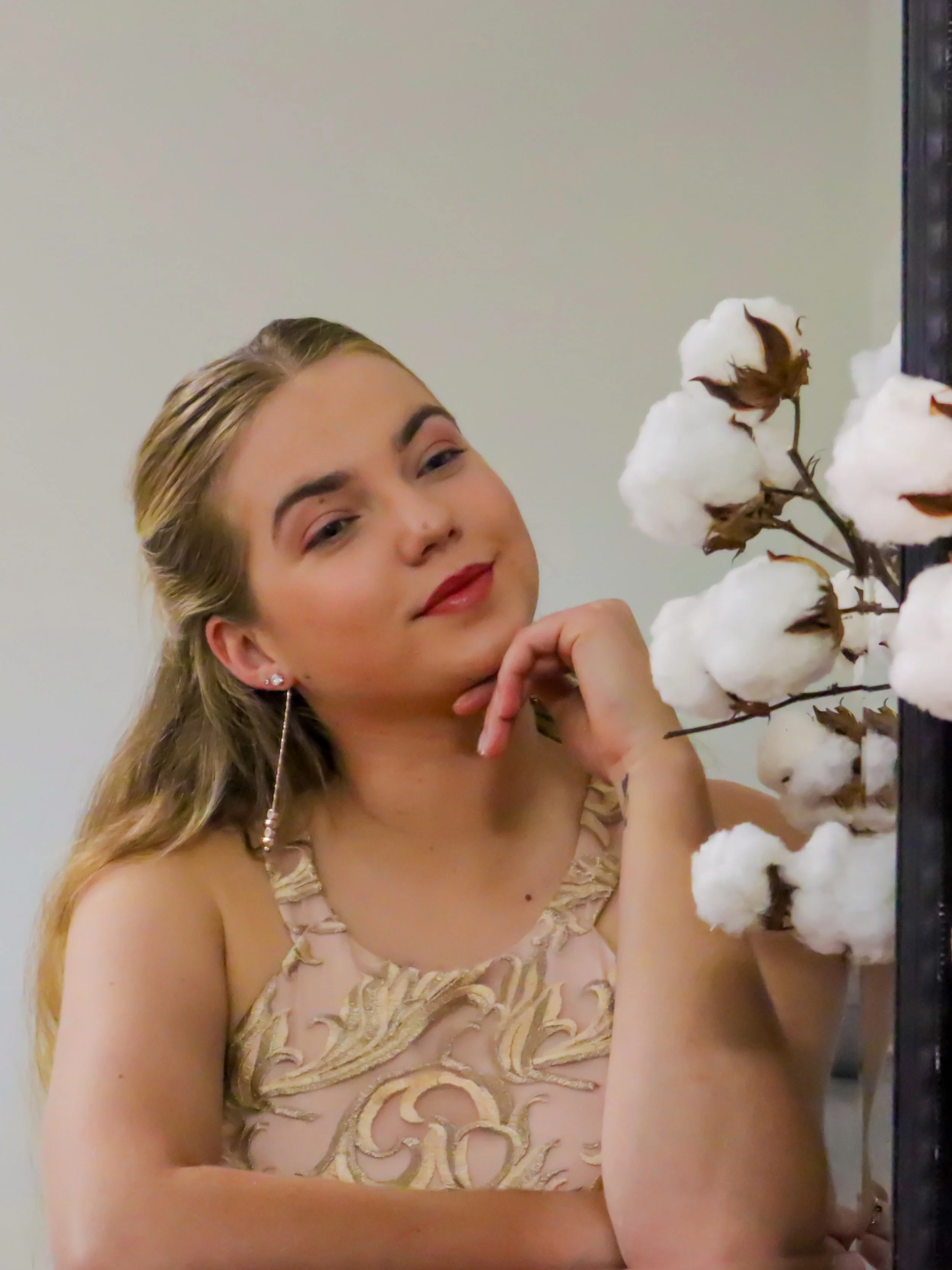A young woman with blonde wavy hair and earrings, wearing a beige dress with gold embroidery, looks at the camera with a slight smile, resting her chin on her hand, next to a branch of white cotton flowers.
