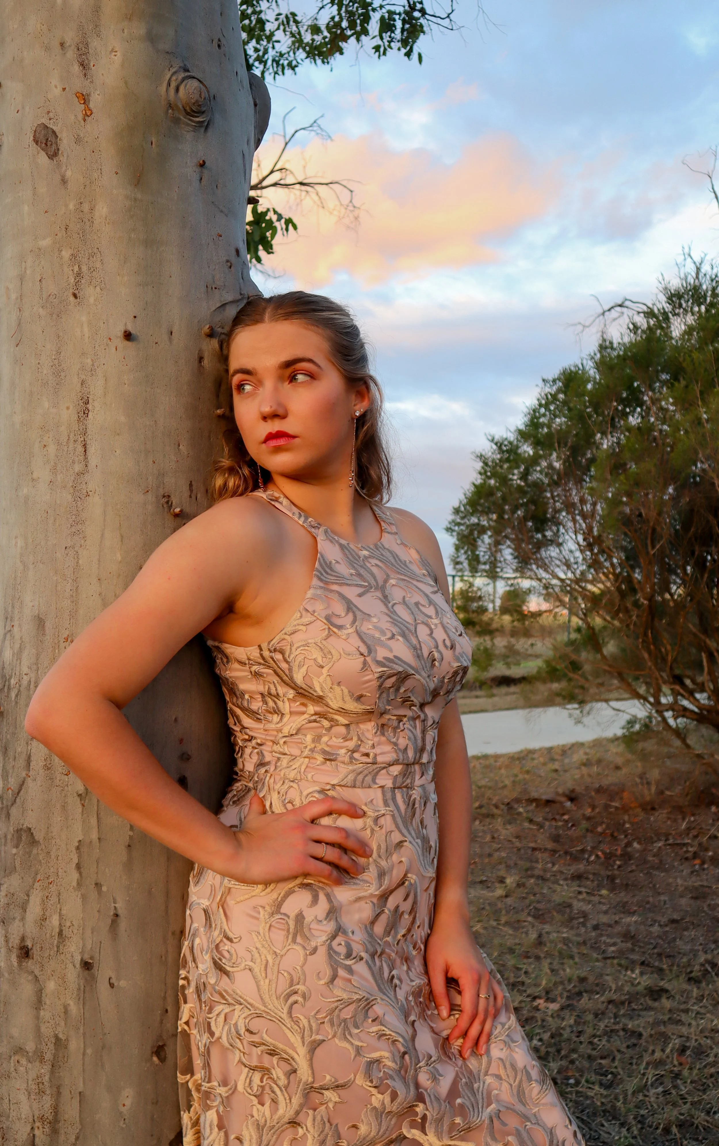 A young woman in a patterned dress leaning against a tree outdoors during sunset.