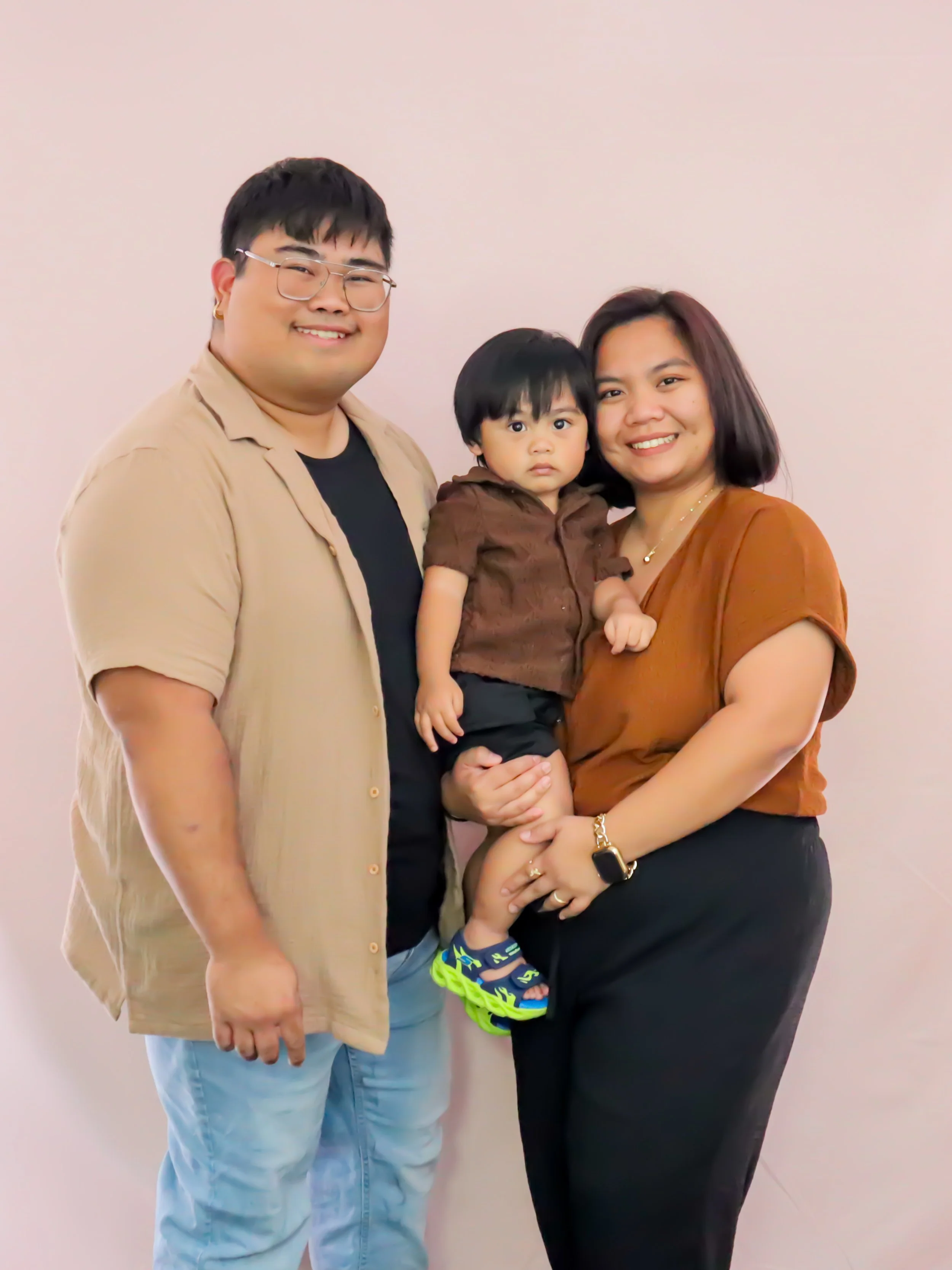 A happy family of three standing together against a plain white wall. The father, wearing glasses and a beige shirt, smiles. The mother, in a brown top, holds their young son, who is dressed in a brown shirt and black shorts, with bright neon sandals