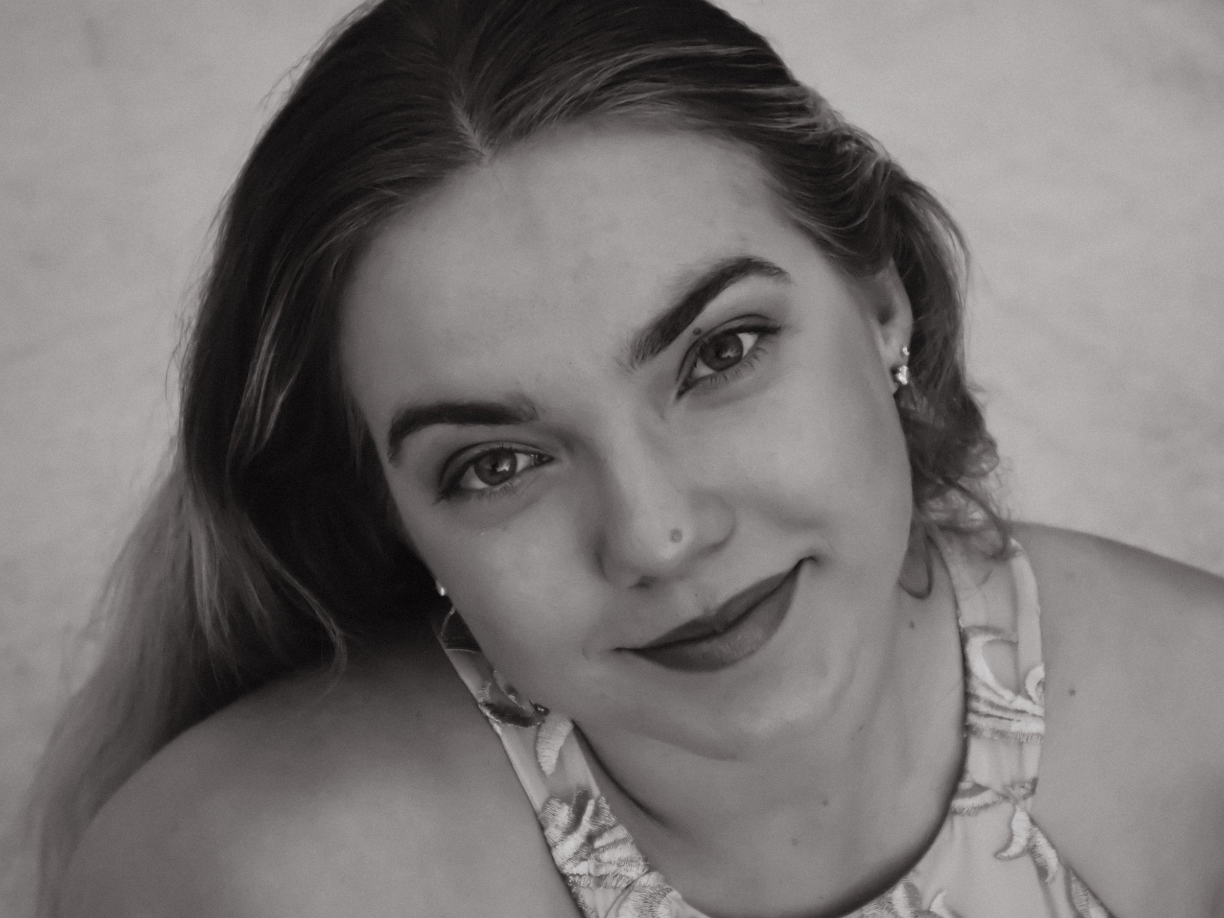 Black and white portrait of a young woman with long hair, wearing earrings, a nostril piercing, and a patterned sleeveless top, smiling slightly at the camera.
