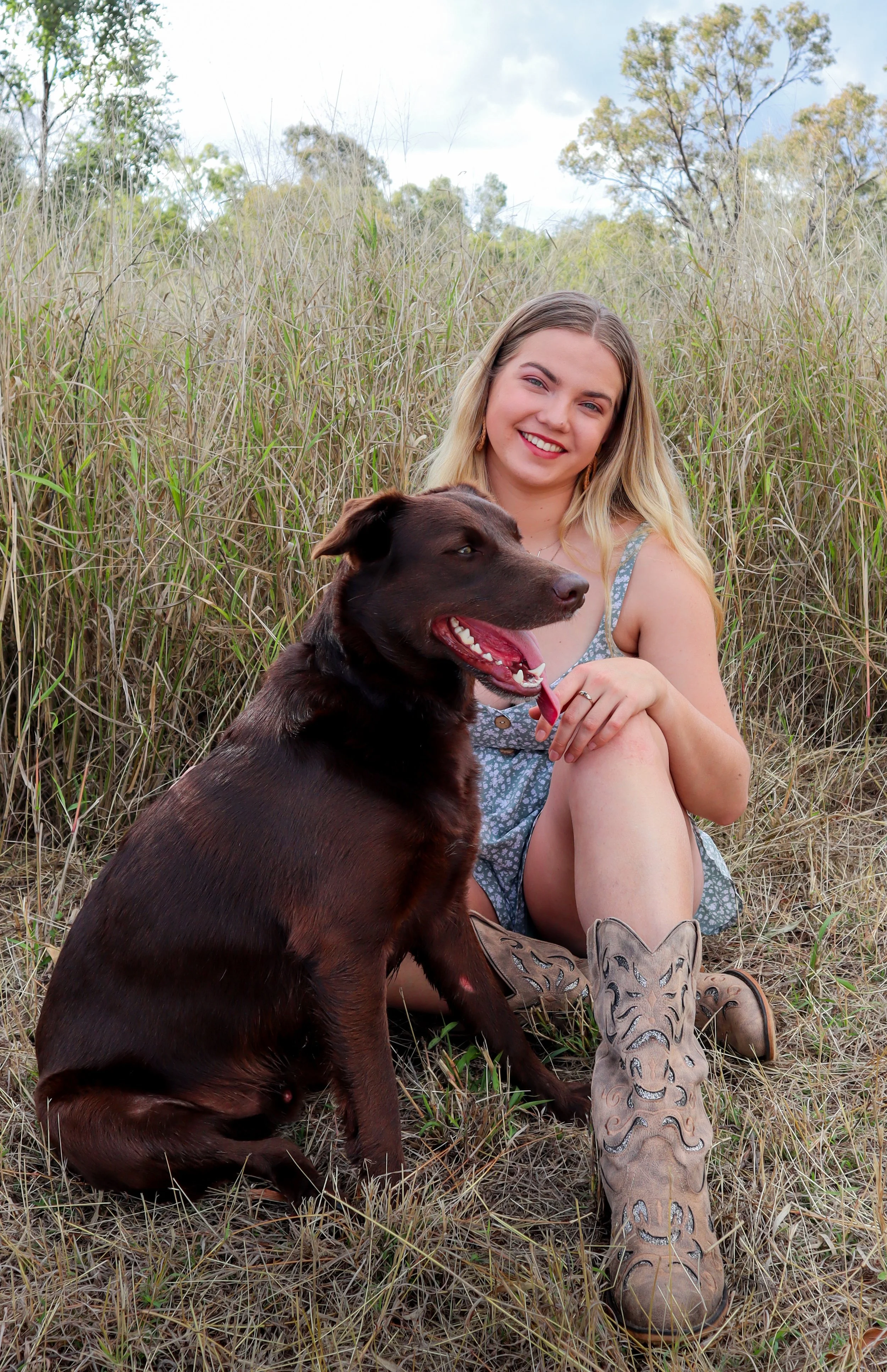 A young woman with blonde hair, wearing a blue dress and cowboy boots, sitting in a grassy field with a large brown dog sitting beside her. The woman is smiling.