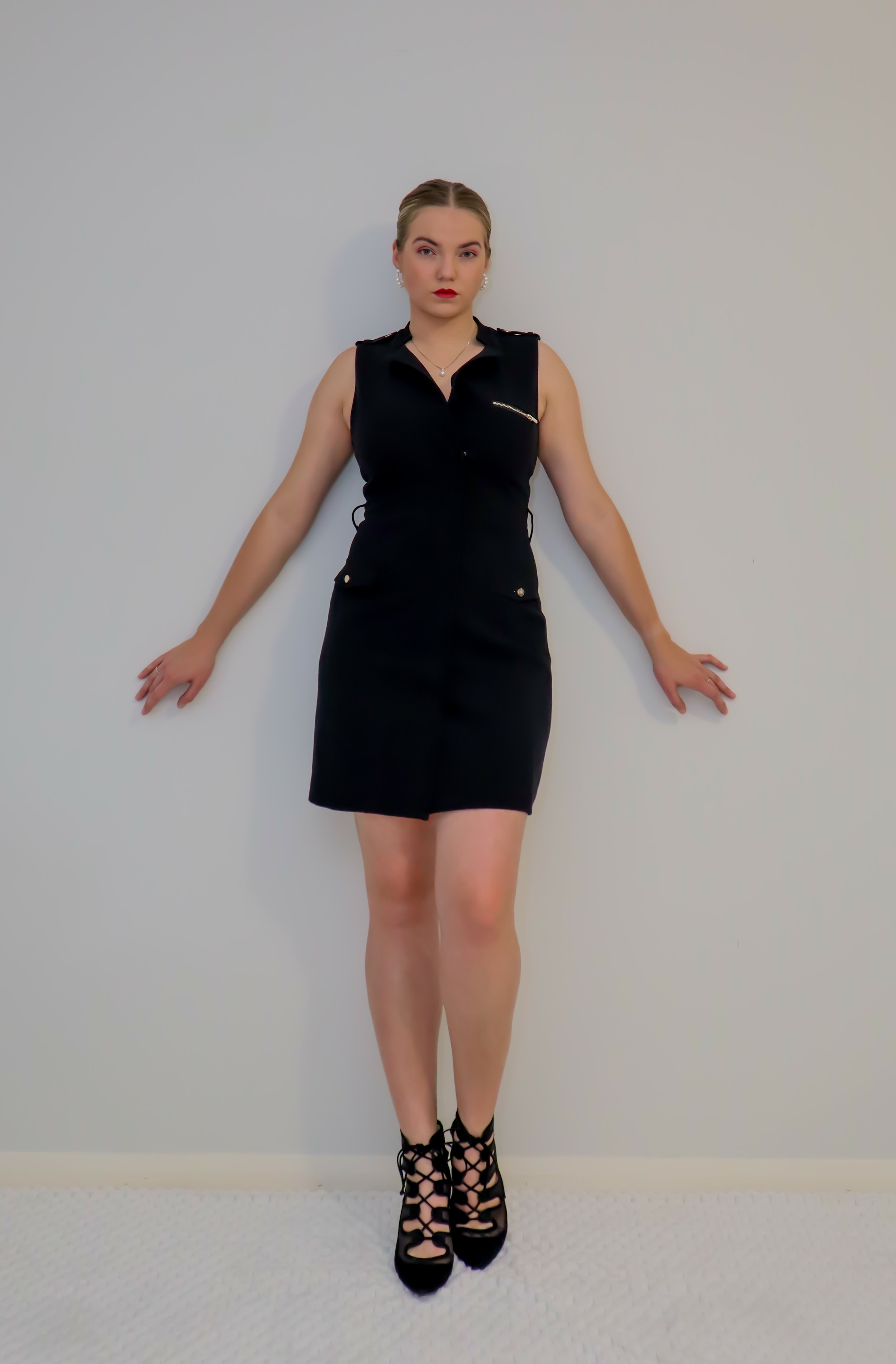 A woman with blonde hair styled in an updo, wearing makeup with lipstick, a black sleeveless dress with gold buttons and a zip pocket, black high heels with lace-up design, standing against a plain white wall.