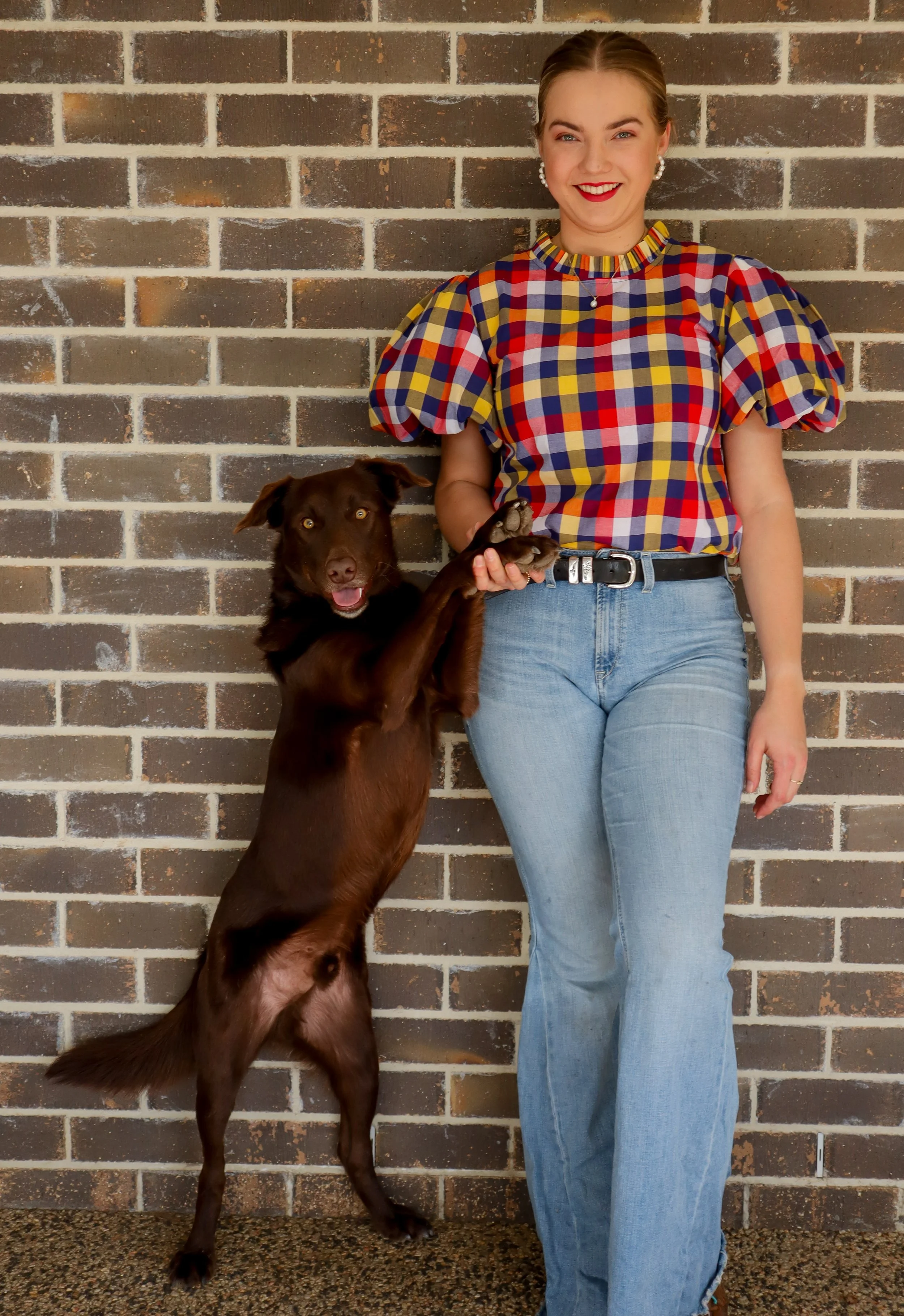 A woman with brown hair in a bun, wearing a colorful plaid shirt, light blue jeans, and red lipstick, standing next to a brown dog with floppy ears against a brick wall. The dog is standing on its hind legs with front paws resting on the woman's hand