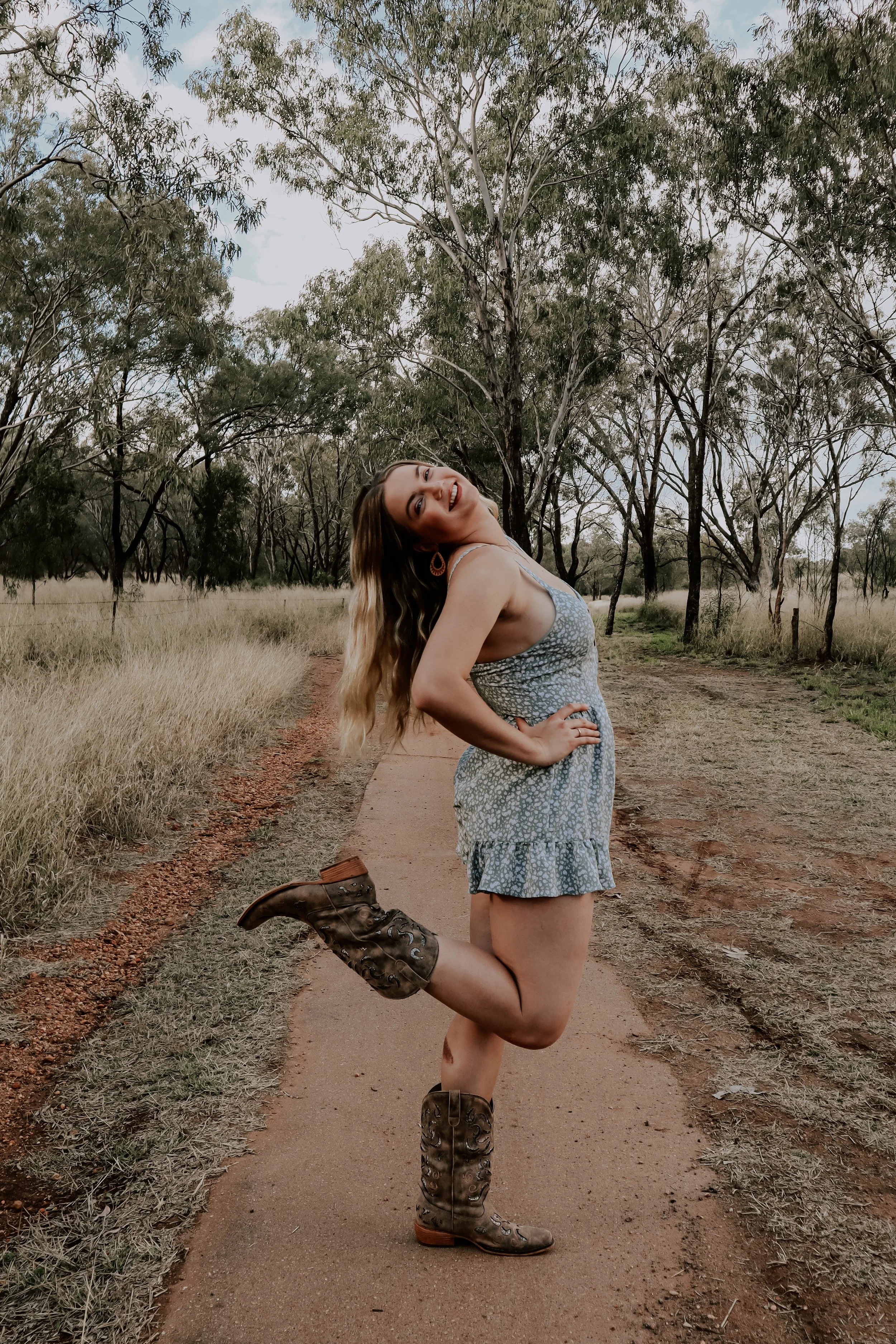 A woman standing on a dirt path in a rural area with trees, wearing a light blue dress and cowboy boots, smiling and posing with one leg bent behind her.