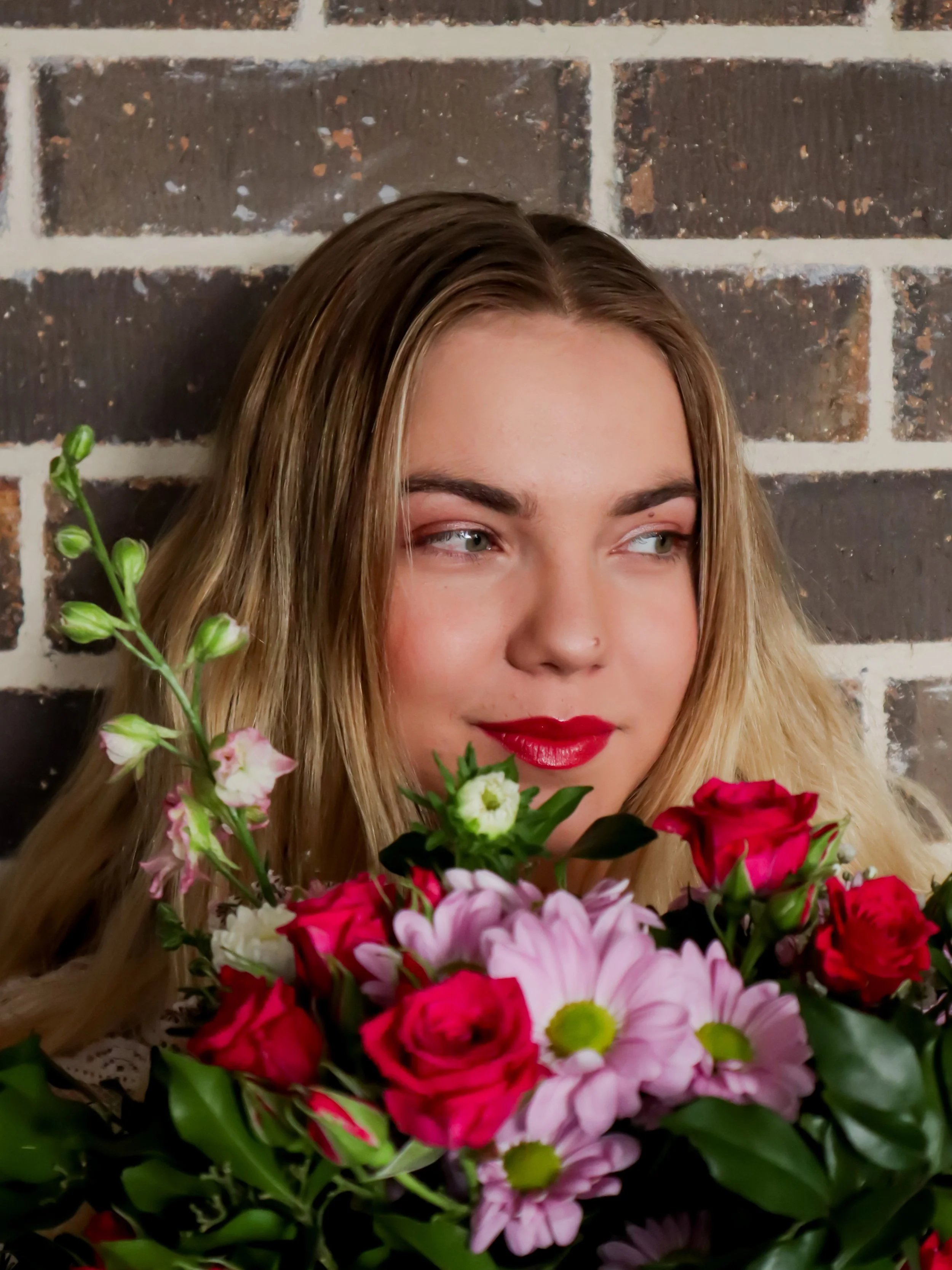 A young woman with blonde hair and red lipstick holding a bouquet of pink, red, and white flowers in front of a brick wall.