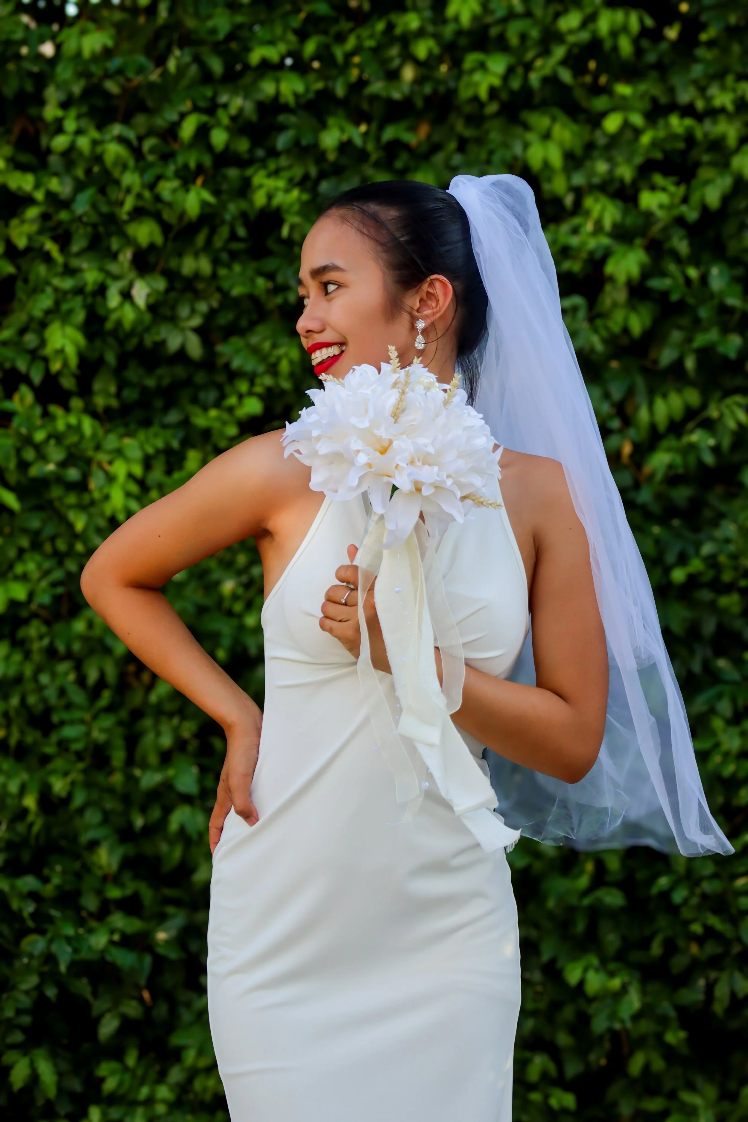 A smiling woman in a white wedding dress holding a bouquet of white flowers, wearing a veil, with a green leafy background.