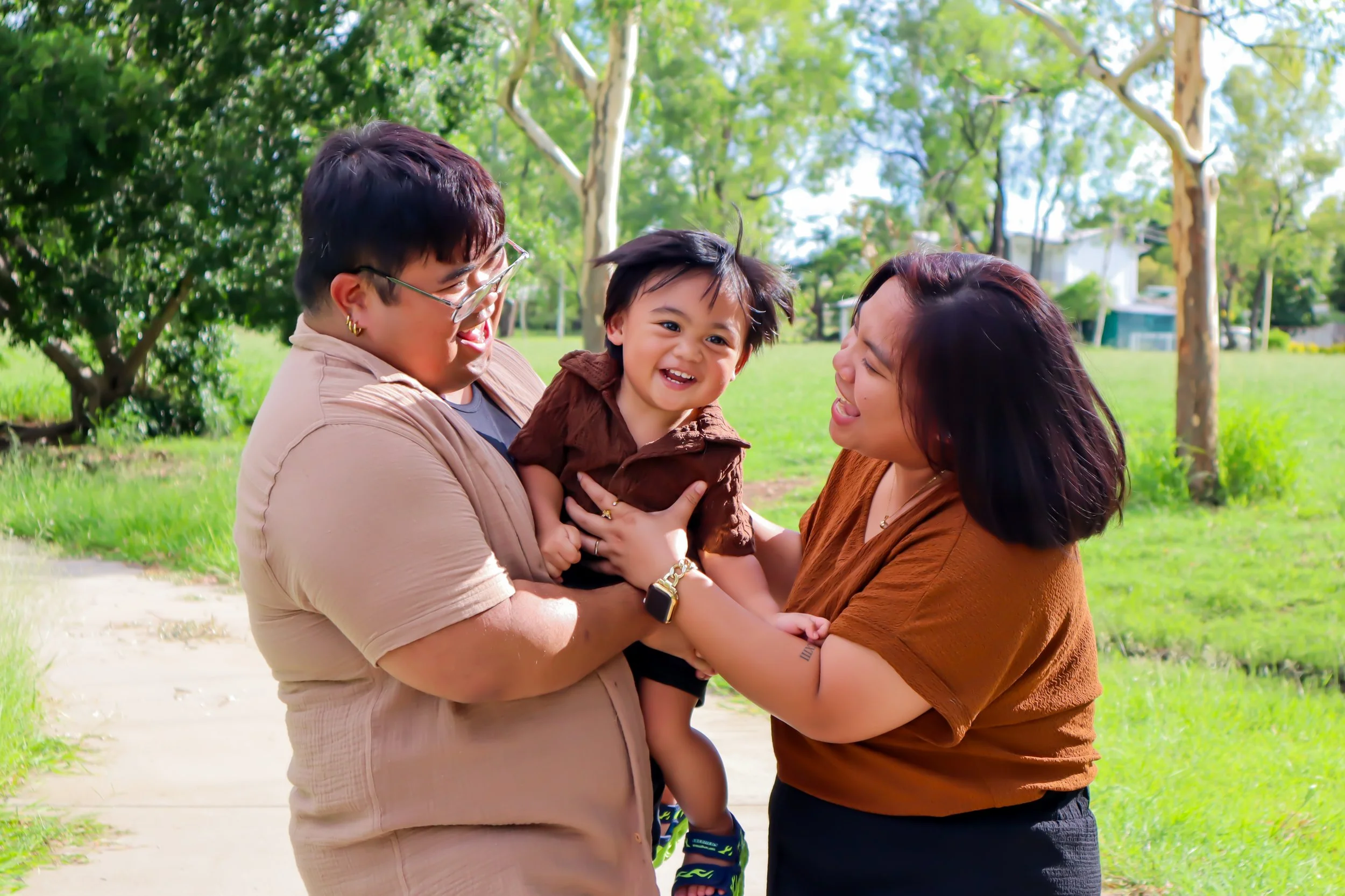 A smiling family of three, including a man, woman, and young child, enjoying a sunny day outdoors in a park. The man and woman are holding the child between them, all laughing and interacting happily.