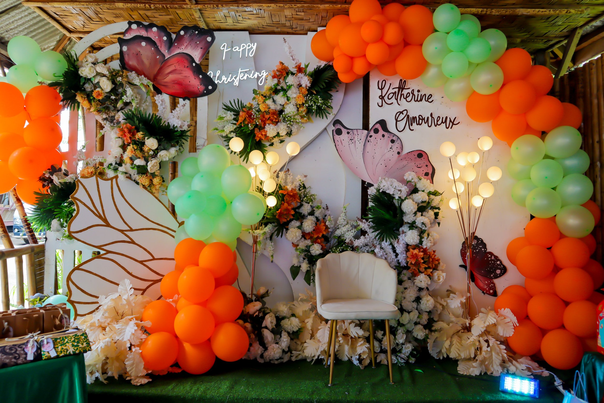 Decorative backdrop with white chair, floral arrangements, butterfly illustrations, and colorful balloons in green and orange for a celebration with the messages 'Happy Christening' and 'Katherine Amoureux'.
