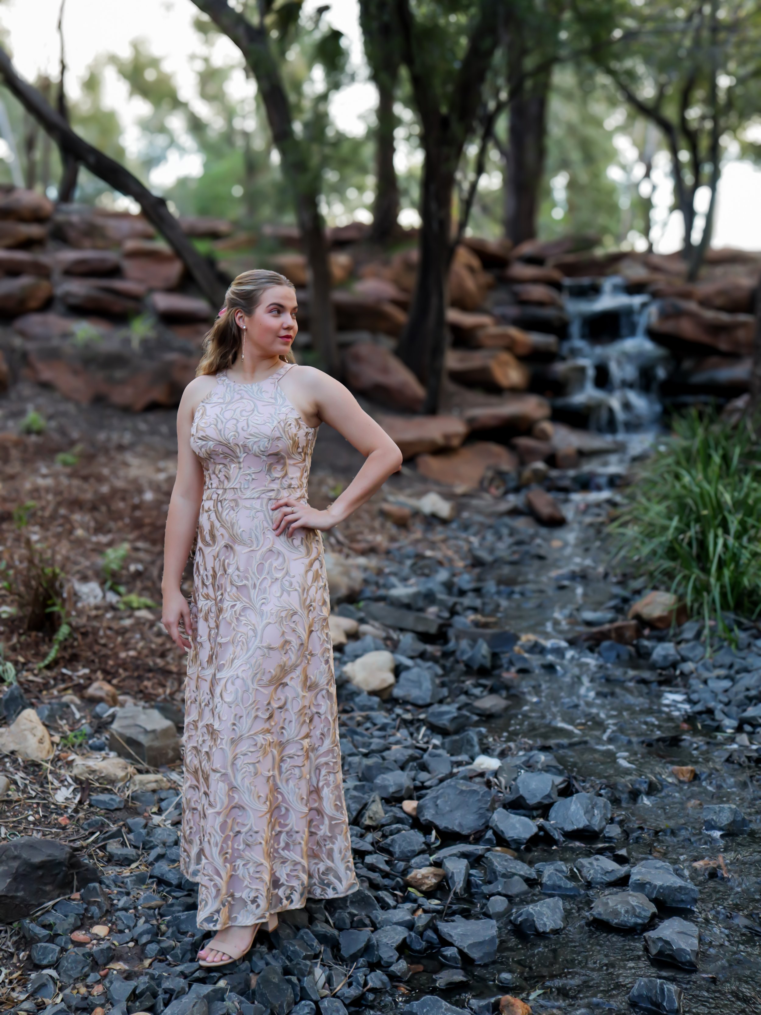 A woman in a beige and gold embroidered dress standing on rocky ground near a small waterfall and stream in a wooded area.