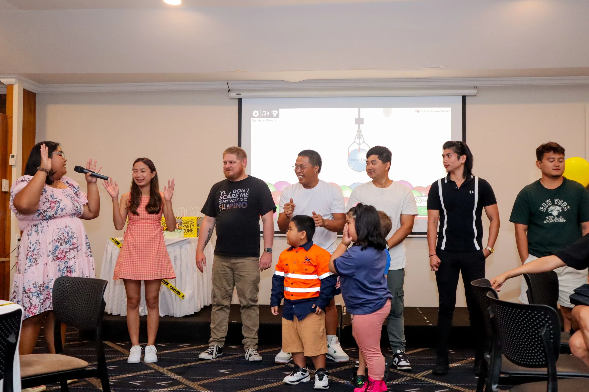 A group of people standing in front of a screen, participating in a game or activity, with a woman holding a microphone and speaking to the group.
