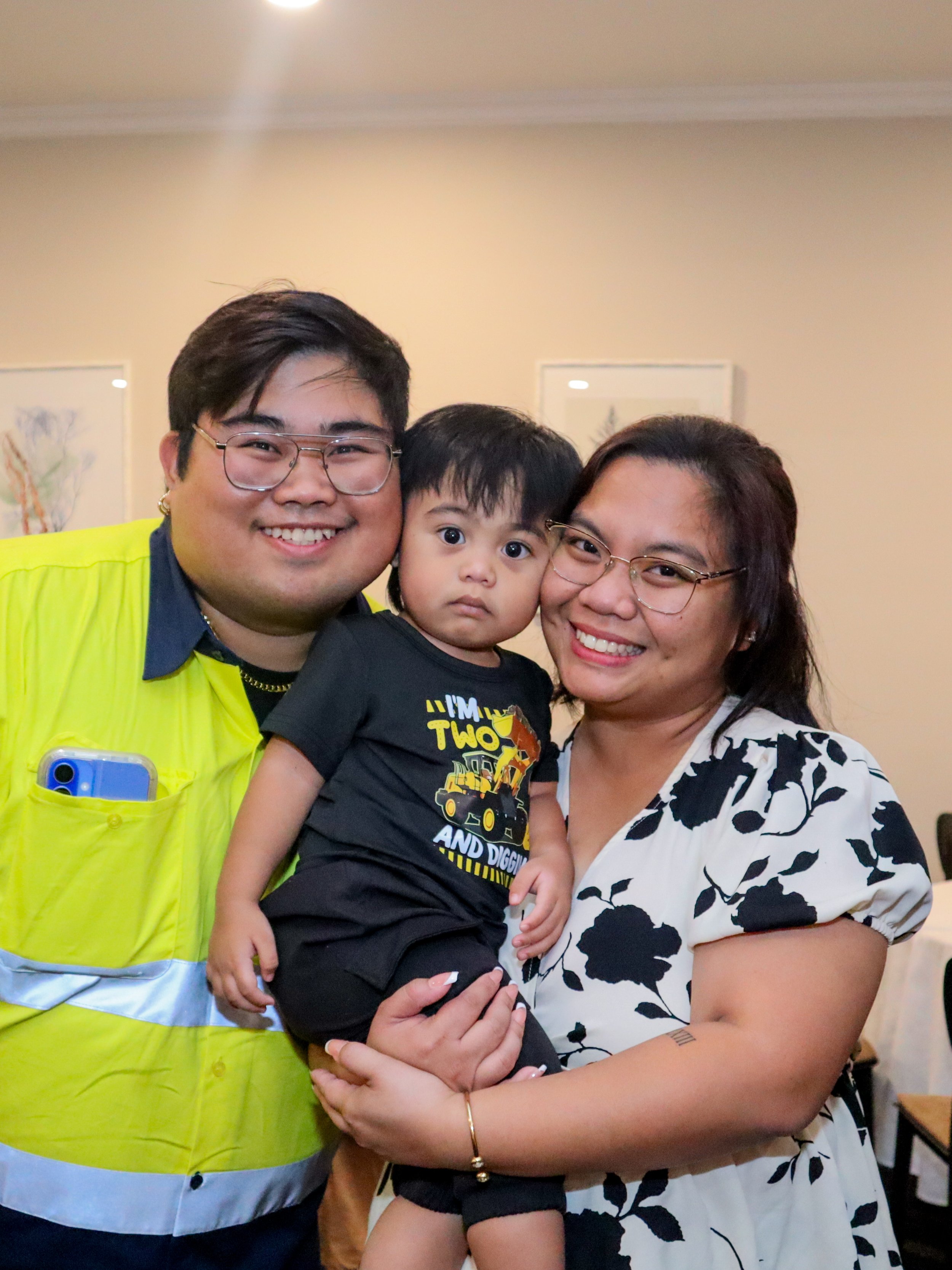 Three people, two adults and a young child, smiling and posing together indoors. The man on the left is wearing glasses and a bright yellow safety vest, and the woman on the right has glasses and a black and white floral dress. The child in the middl