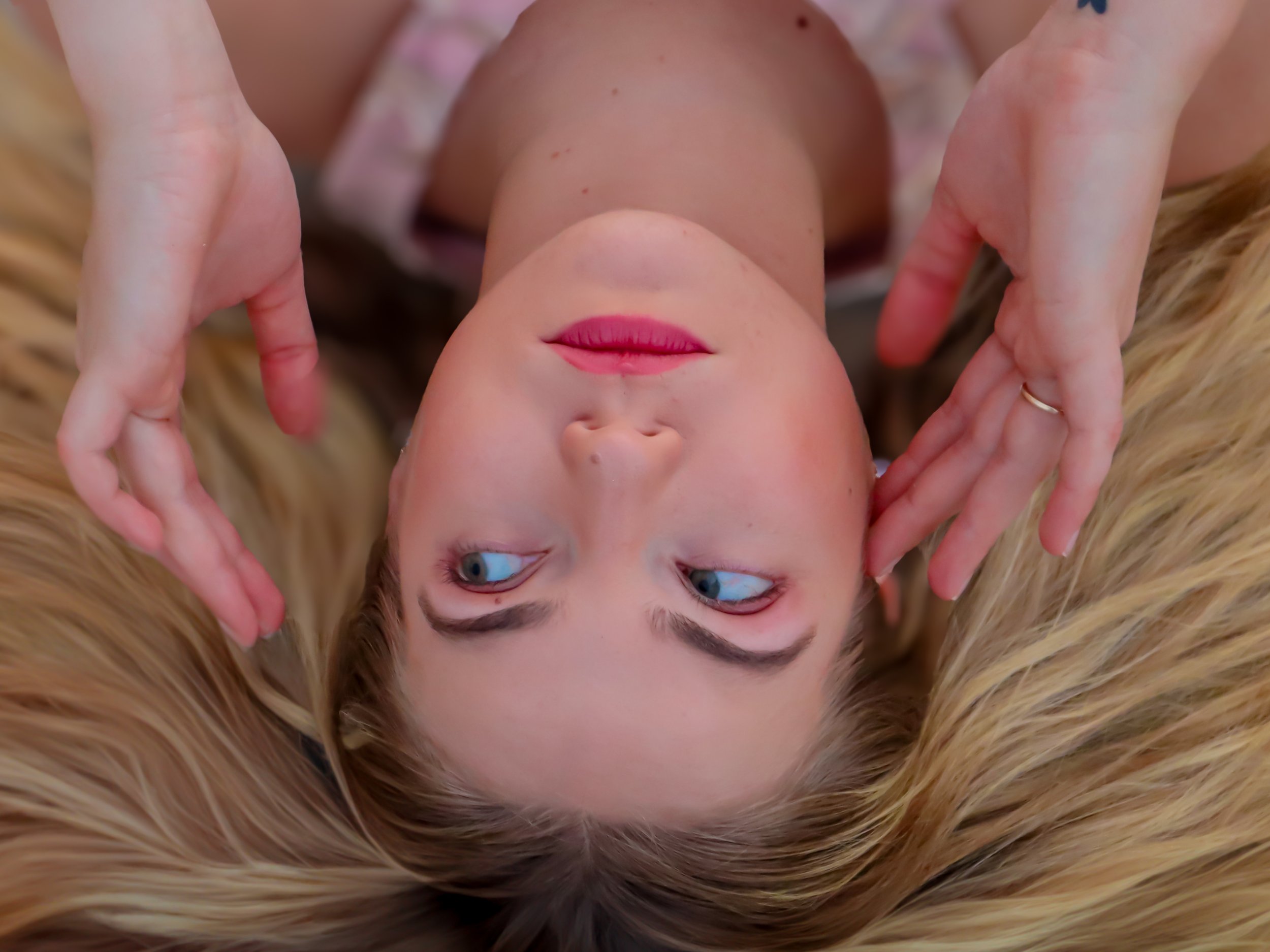 A woman lying down with her head surrounded by long blonde hair, looking sideways with her hands framing her face, wearing pink lipstick and having bright blue eyes.