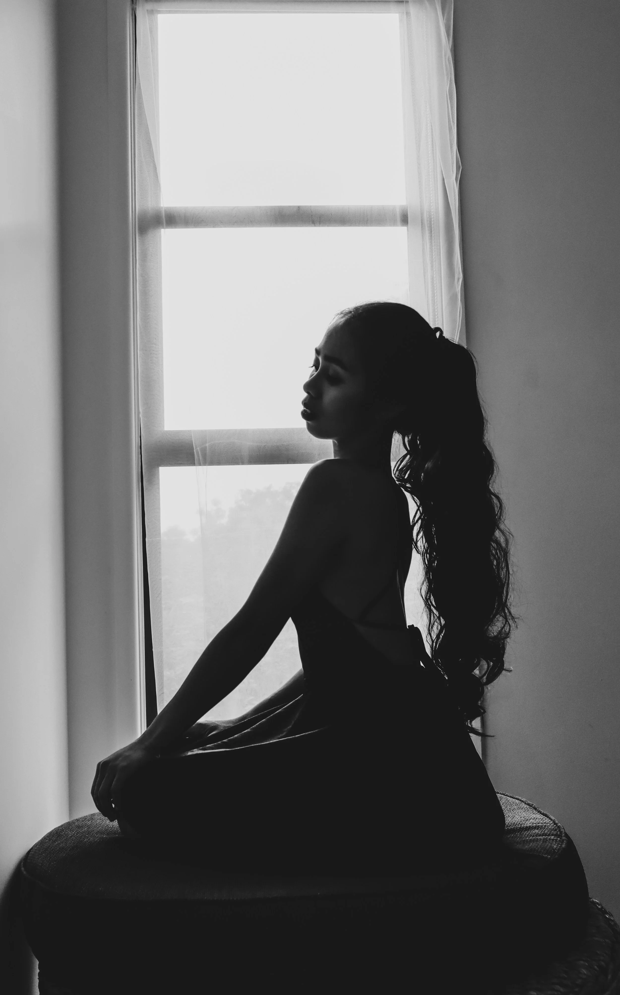 A black and white silhouette of a woman sitting on a cushioned surface near a window, with her eyes closed and long, wavy hair flowing down her back.