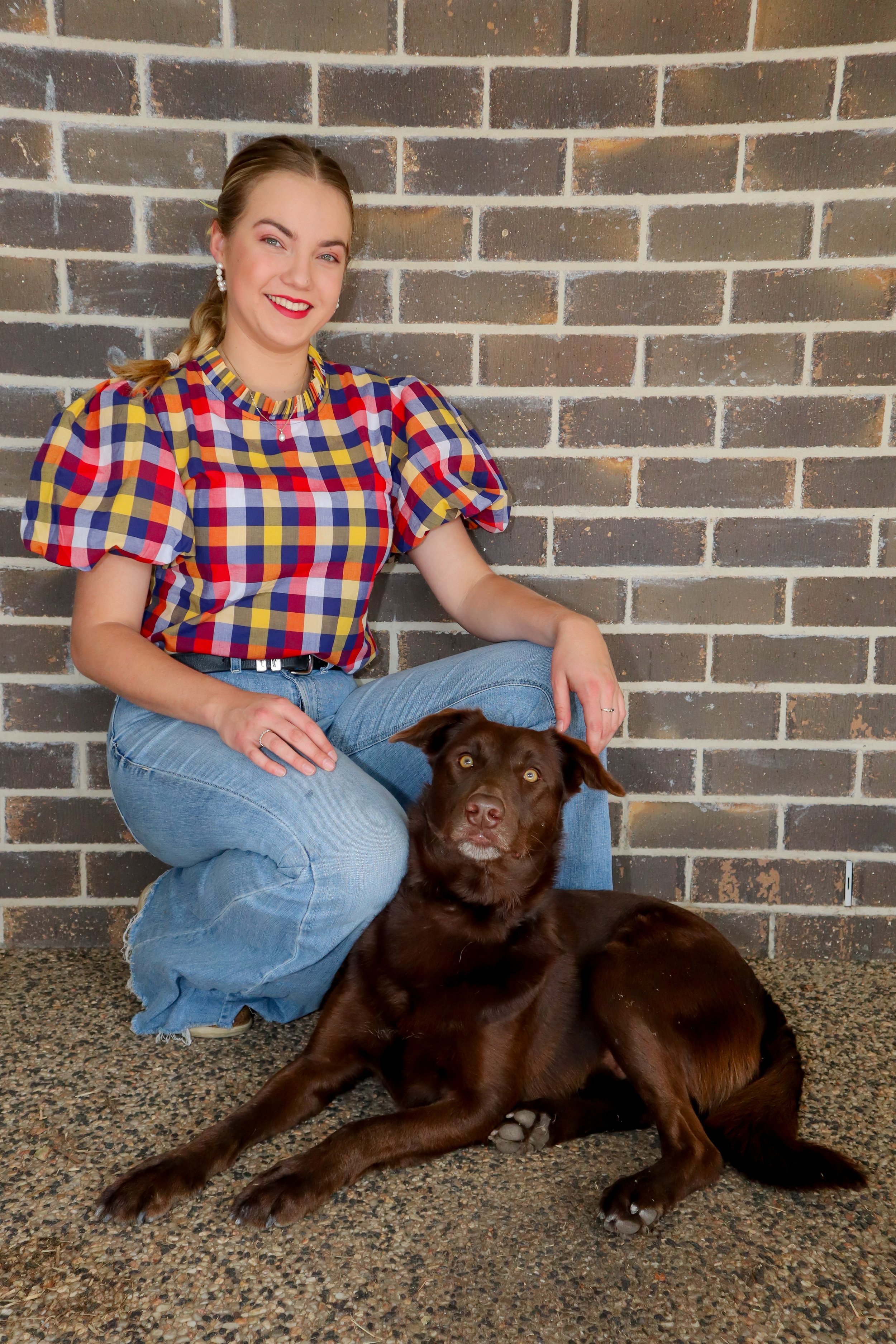 A young woman with a braid wearing a colorful checkered puff-sleeve shirt and light blue jeans is sitting on a speckled floor next to a brown dog with golden eyes, against a brick wall background.