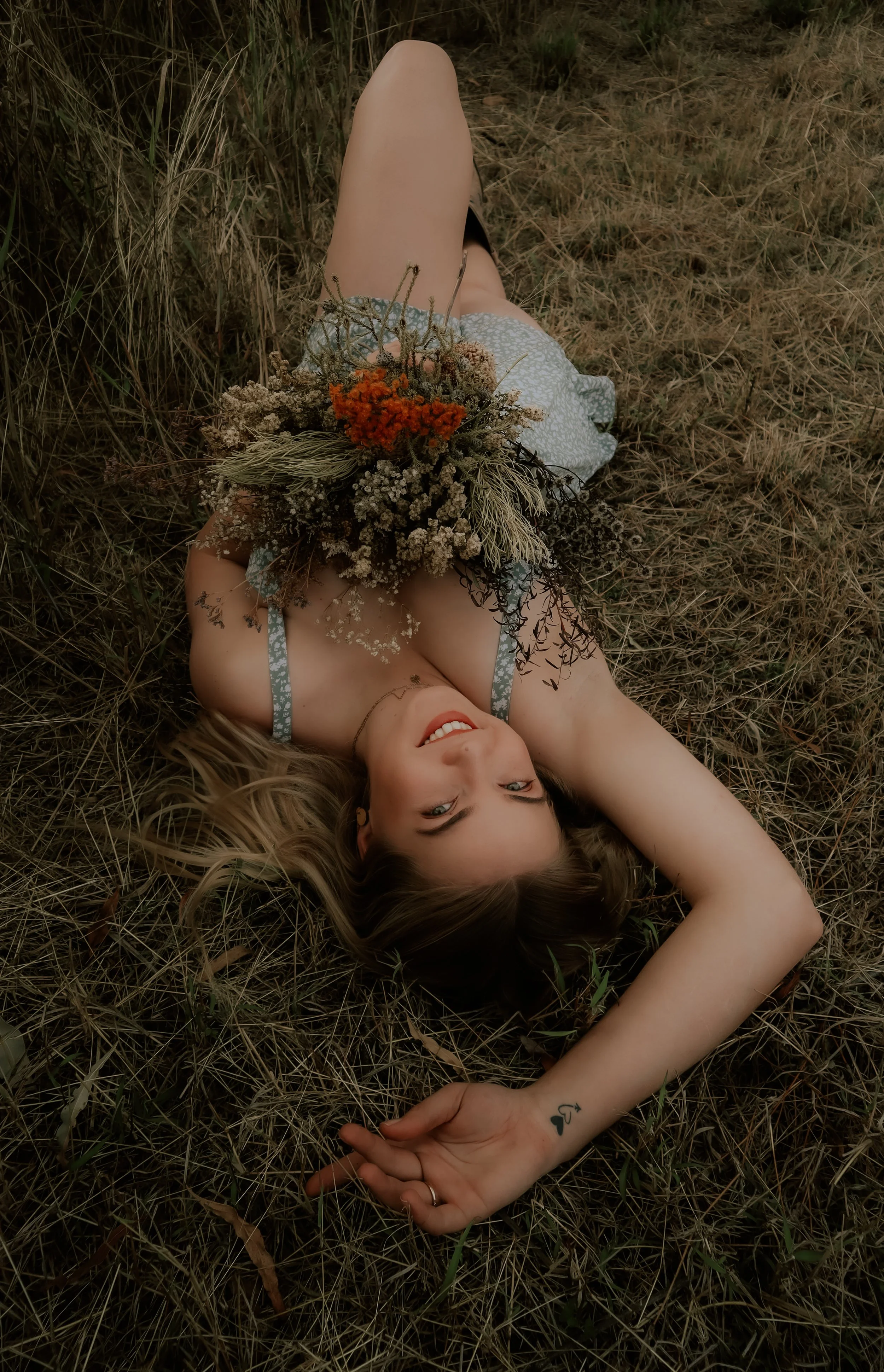 A woman lying on her back in a grassy field, holding a bouquet of wildflowers.