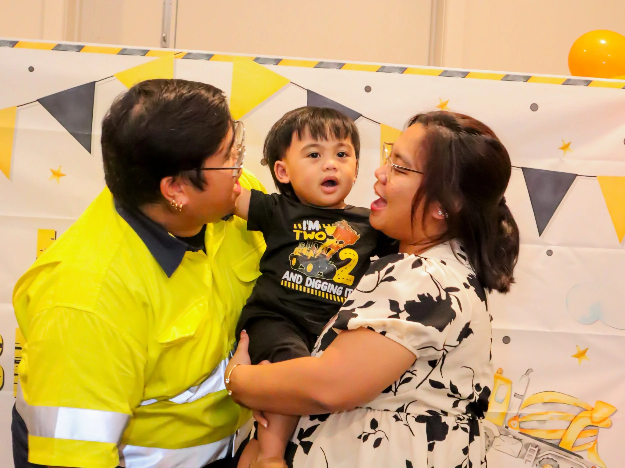 A boy being held by a woman, smiling, with a man holding and smiling at him, at a birthday celebration with yellow and black decorations.