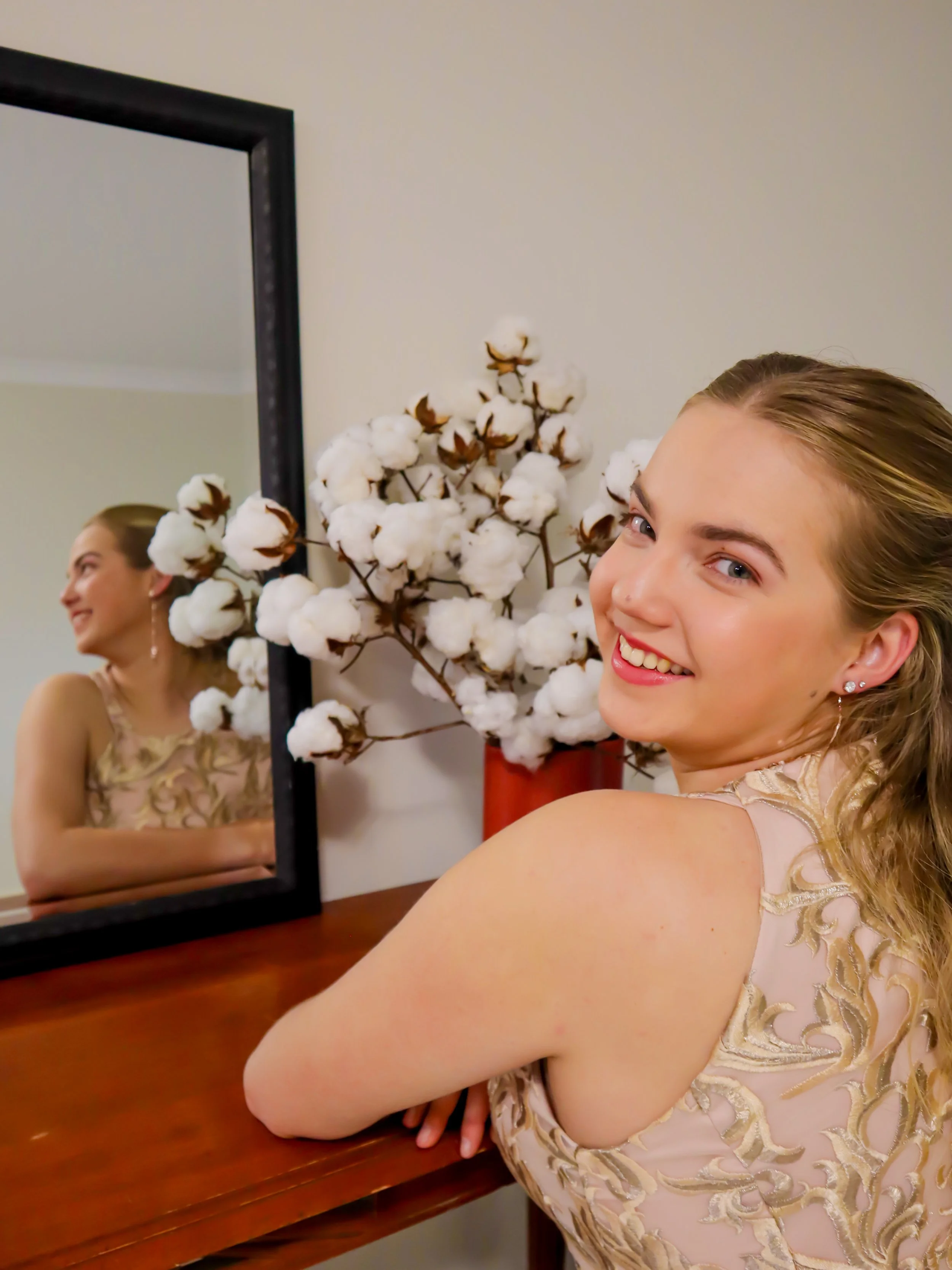 A smiling young woman with blonde hair, wearing a light-colored dress with gold embroidery, is sitting at a wooden table. Behind her, there's a large mirror and a red vase with white cotton branch flowers. The woman's reflection is visible in the mir